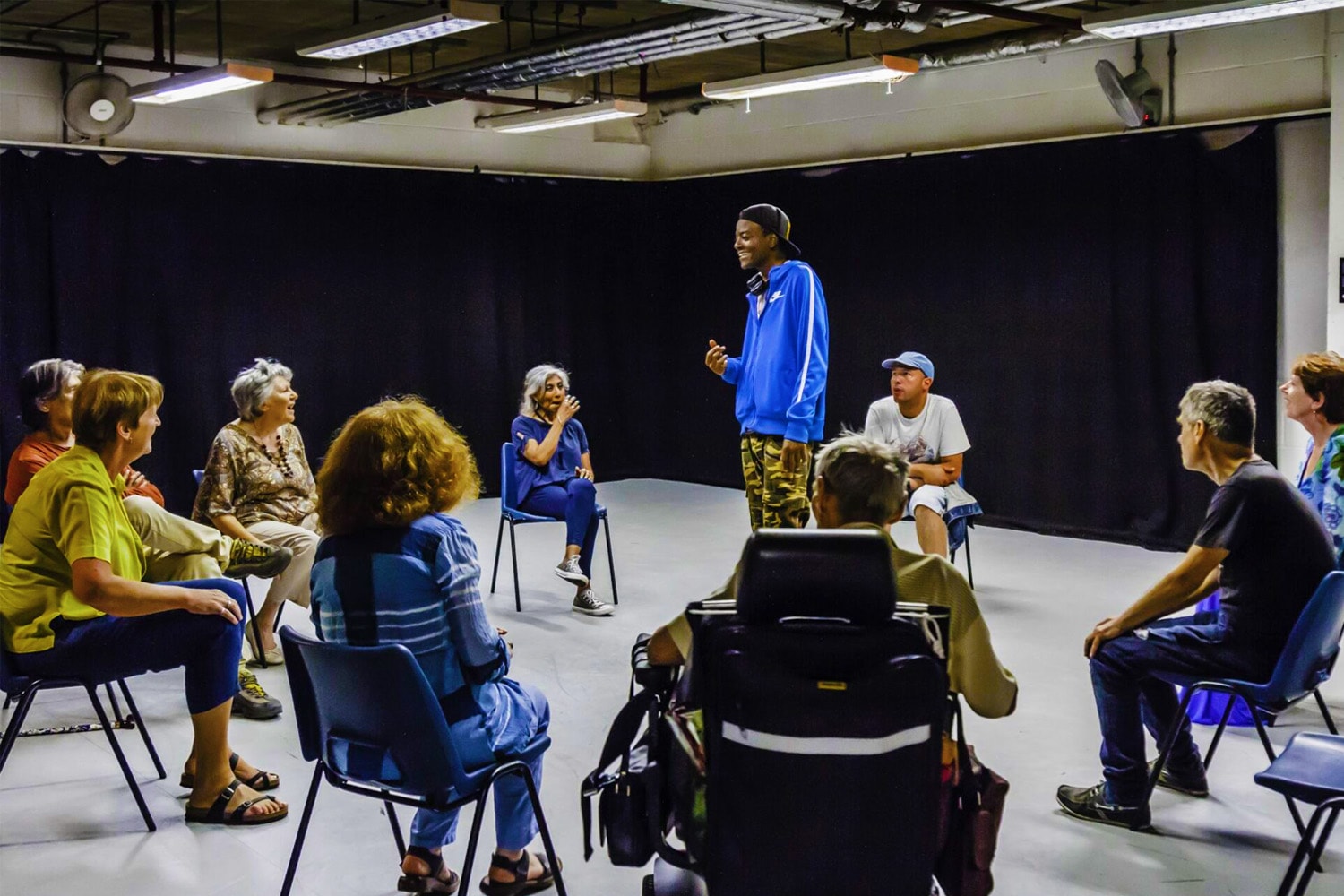 An artsdepot drama workshop in a drama studio. In the centre, a young black man in a blue hoodie is speaking. He is in the middle of a circle of other adults of various ages, all sat down.