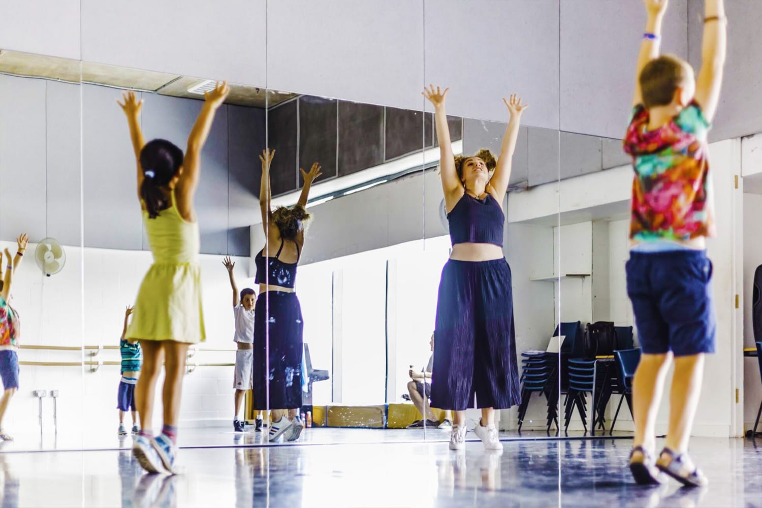 A young person's movement class is being led by an instructor in a dance studio. All participants are stretching upwards towards the ceiling on tiptoes.