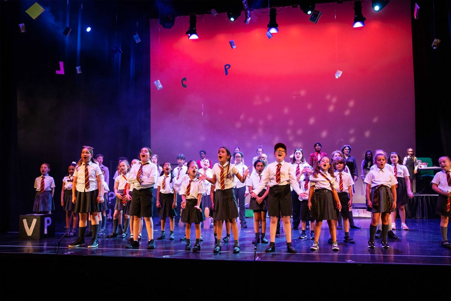 A diverse group of children stand onstage in a group performance of Matilda the musical. They all wear school uniforms with red ties and have their mouths open mid song. Alphabet letters hang down from the ceiling.