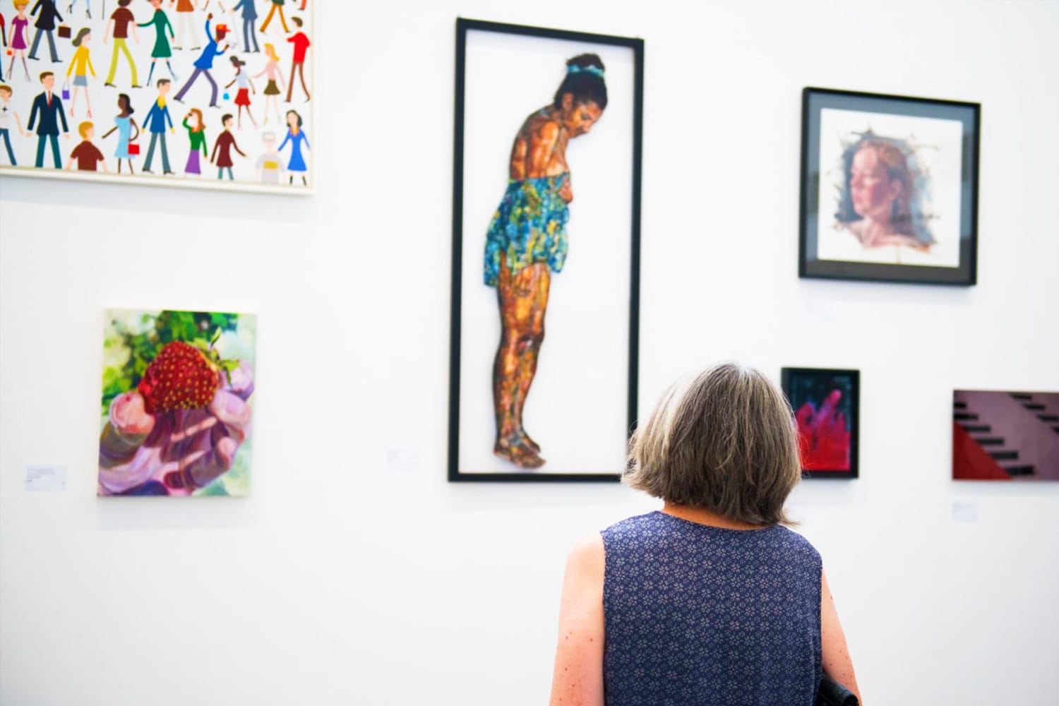 From behind, we see a woman with mid-length grey hair admiring a wall of paintings in an art gallery.