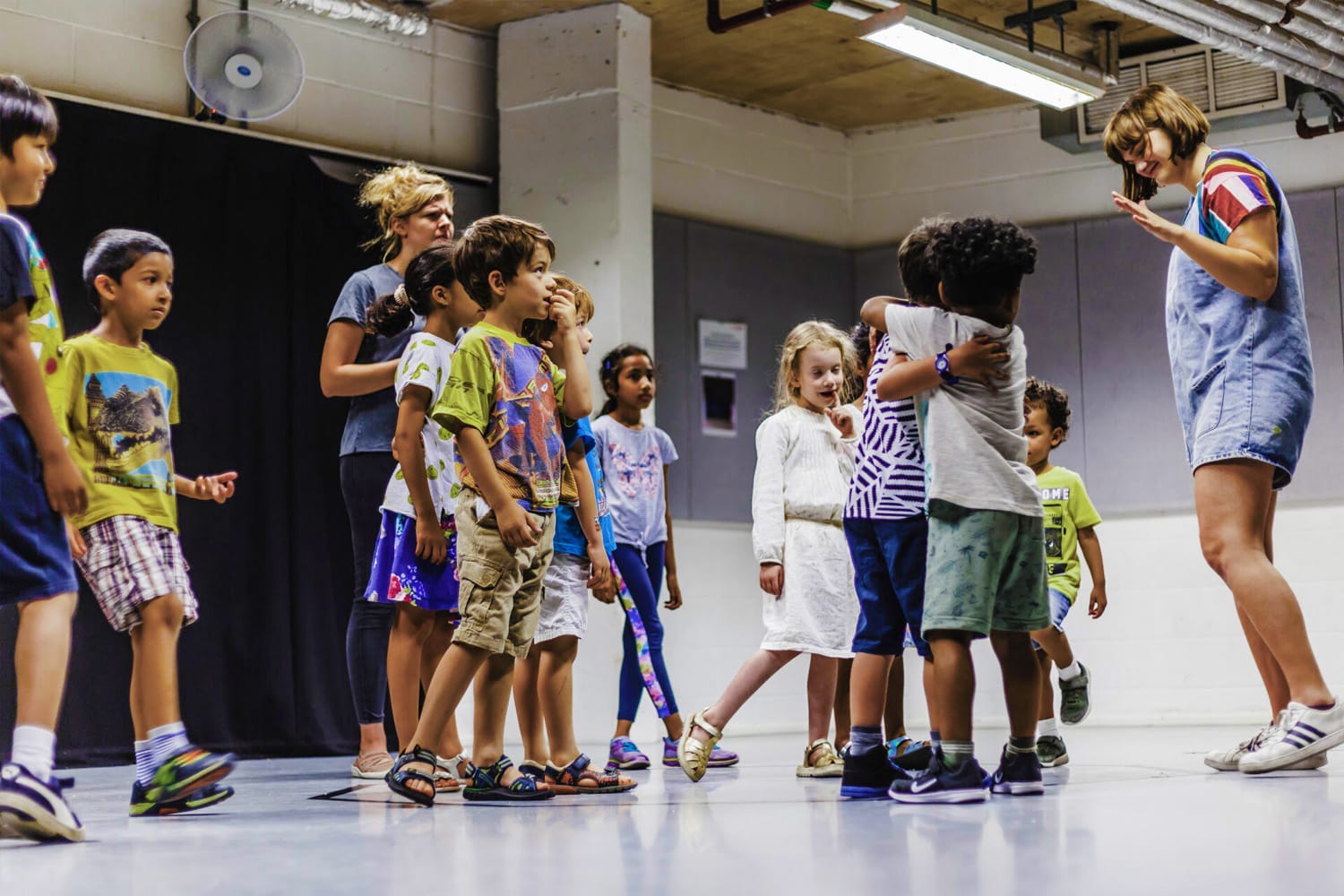 A diverse group of children are engaging in a drama workshop. They all smiling and listening to the group leader. Two boys in the centre of the image are hugging each other. Everyone is wearing colourful clothes and mixed patterns.