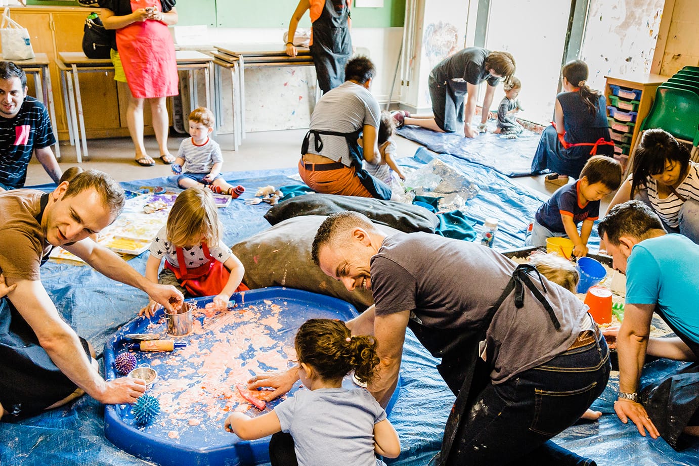 A baby and toddler craft event at artsdepot. Several parents and their young children are engaging in messy sensory play on blue plastic mats. Everyone is smiling and exploring the materials.