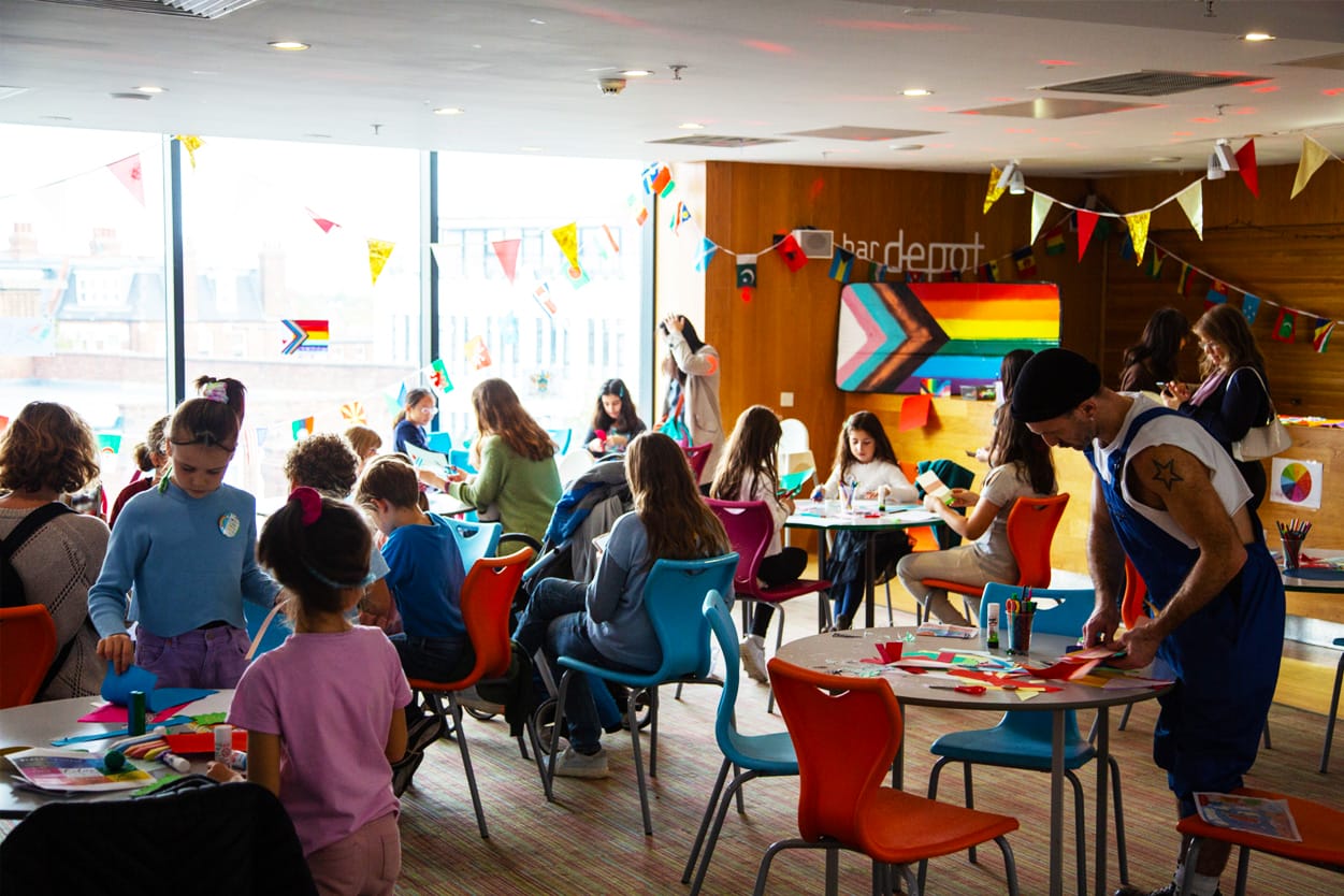 bardepot is filled with colourful chairs and tables for a community event. Children, adults and young people all engage in craft activities. World flags are hung around the windows and a large hand-painted pride flag is featured.