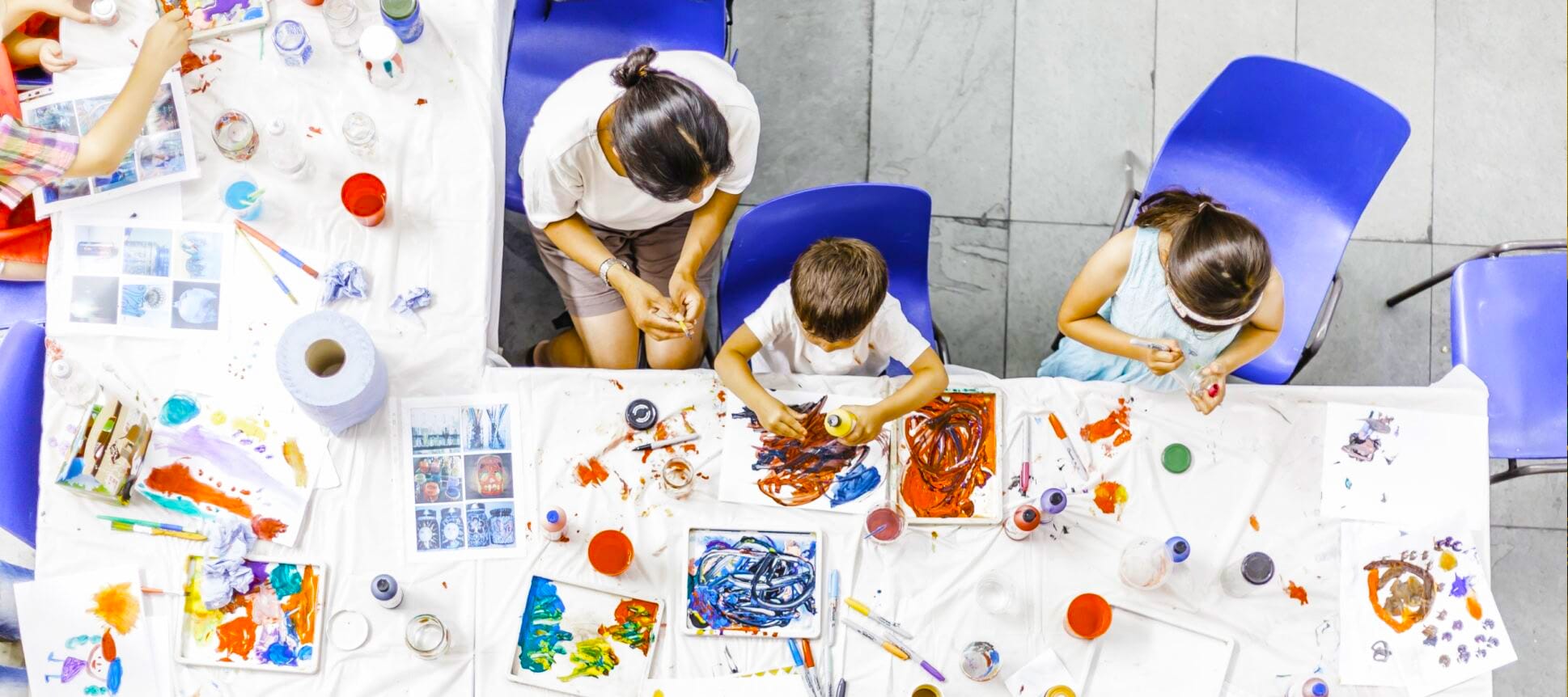 A long, busy table full of messy finger painting, colourful paints and art supplies. An adult participates in the art with two children.