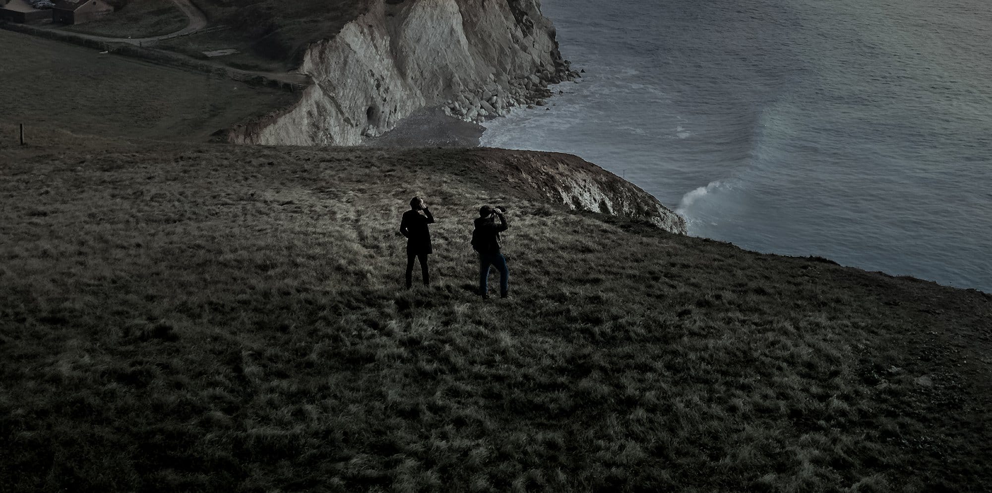 A dark, atmospheric image of a cliffside and coastline. On the land, two silhouette figures look out towards the water.