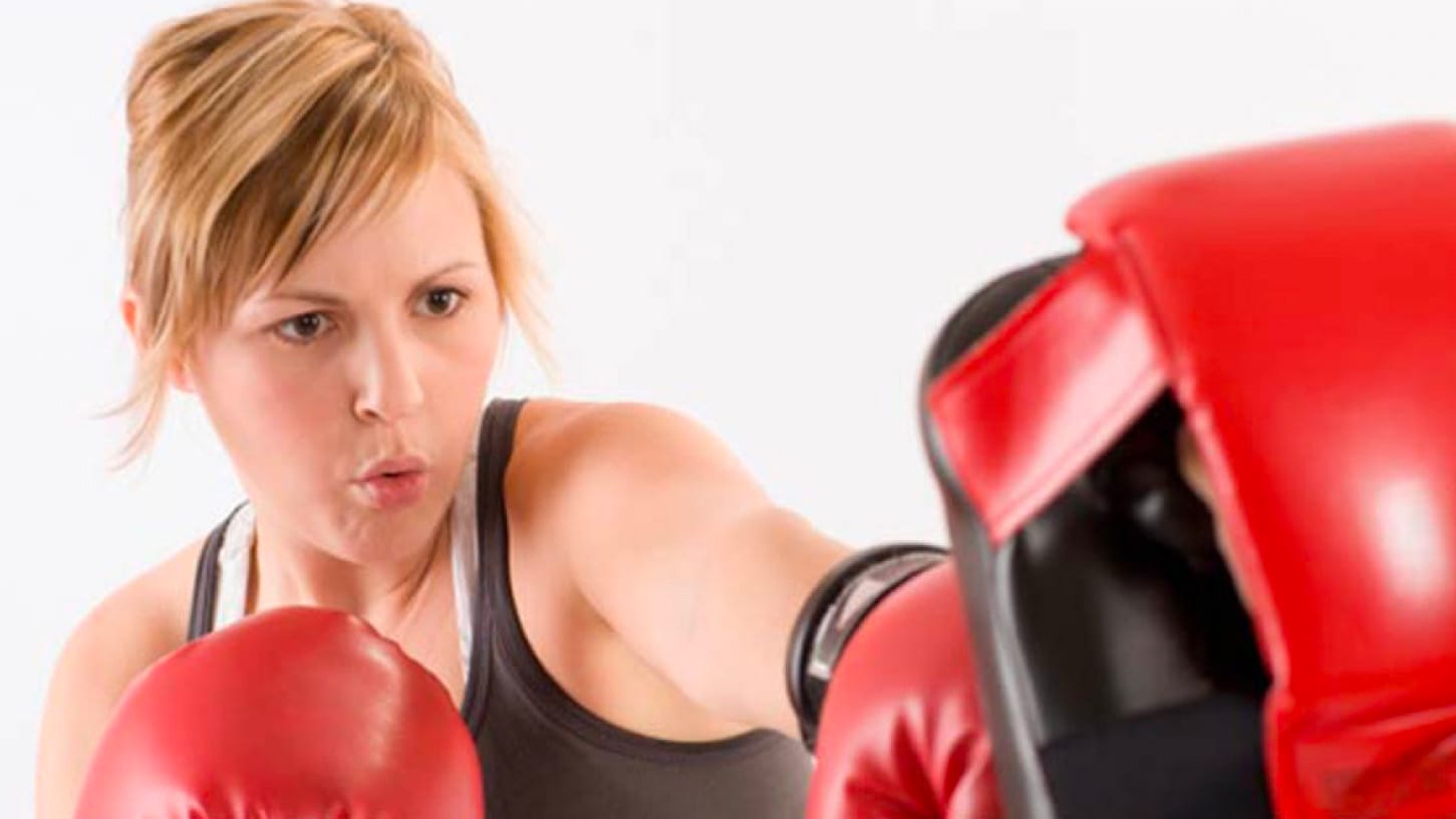 A woman with a blonde ponytail poses mid punch. She sporting wearing red and black boxing gloves.