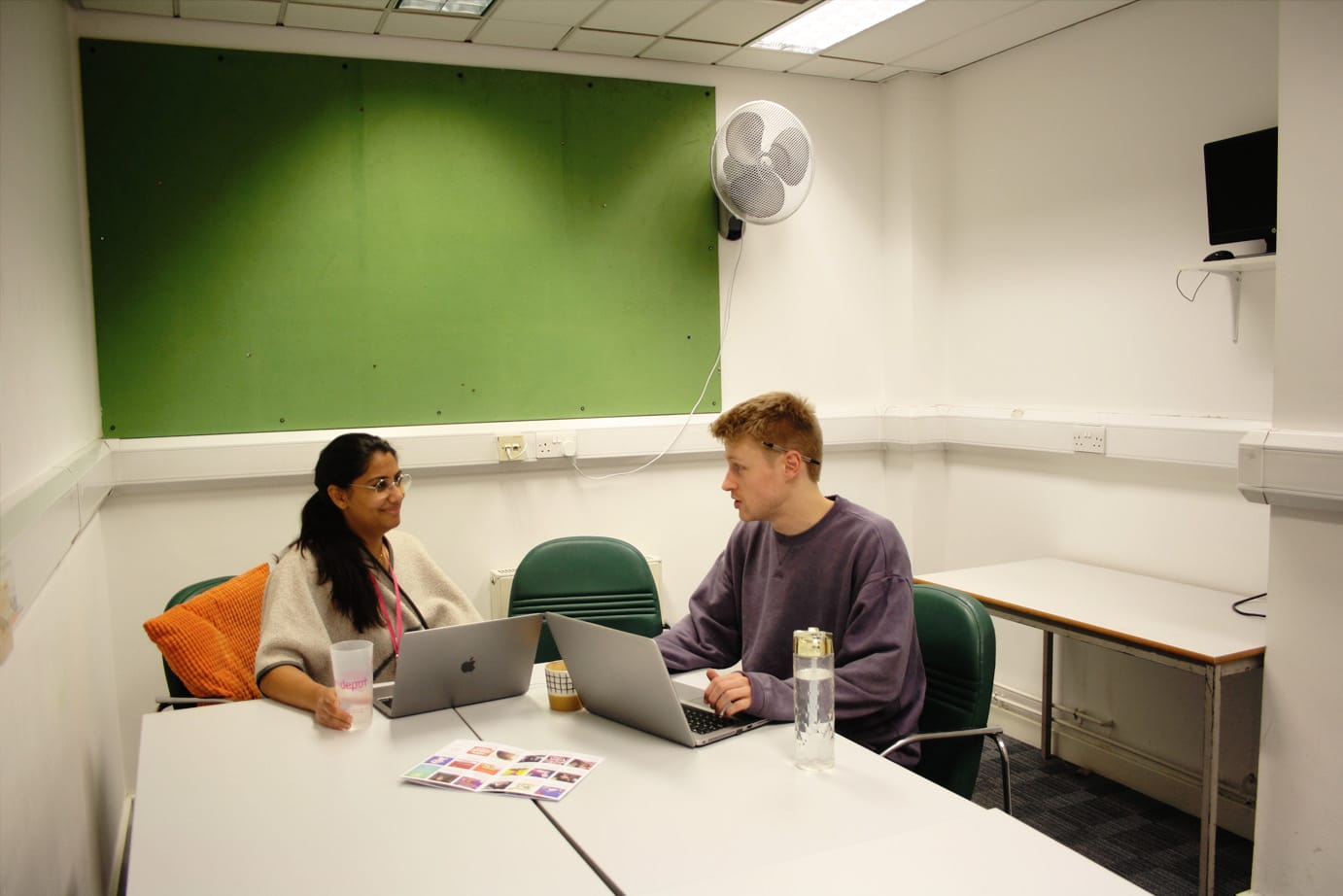 Two artsdepot team members converse in the meeting room. They are engaged in conversation with laptops open and water and coffee cups on the table. Behind them is a green noticeboard.
