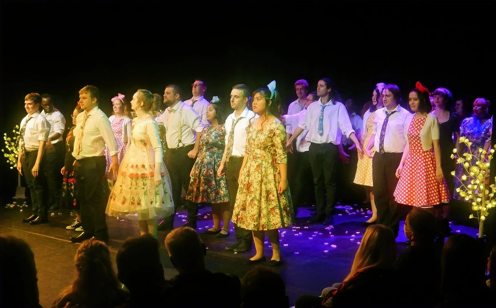 A diverse group of performers photographed on stage in costume. They wear white shorts with colourful ties, and vibrant floral dresses. There are petals scattered across the stage.