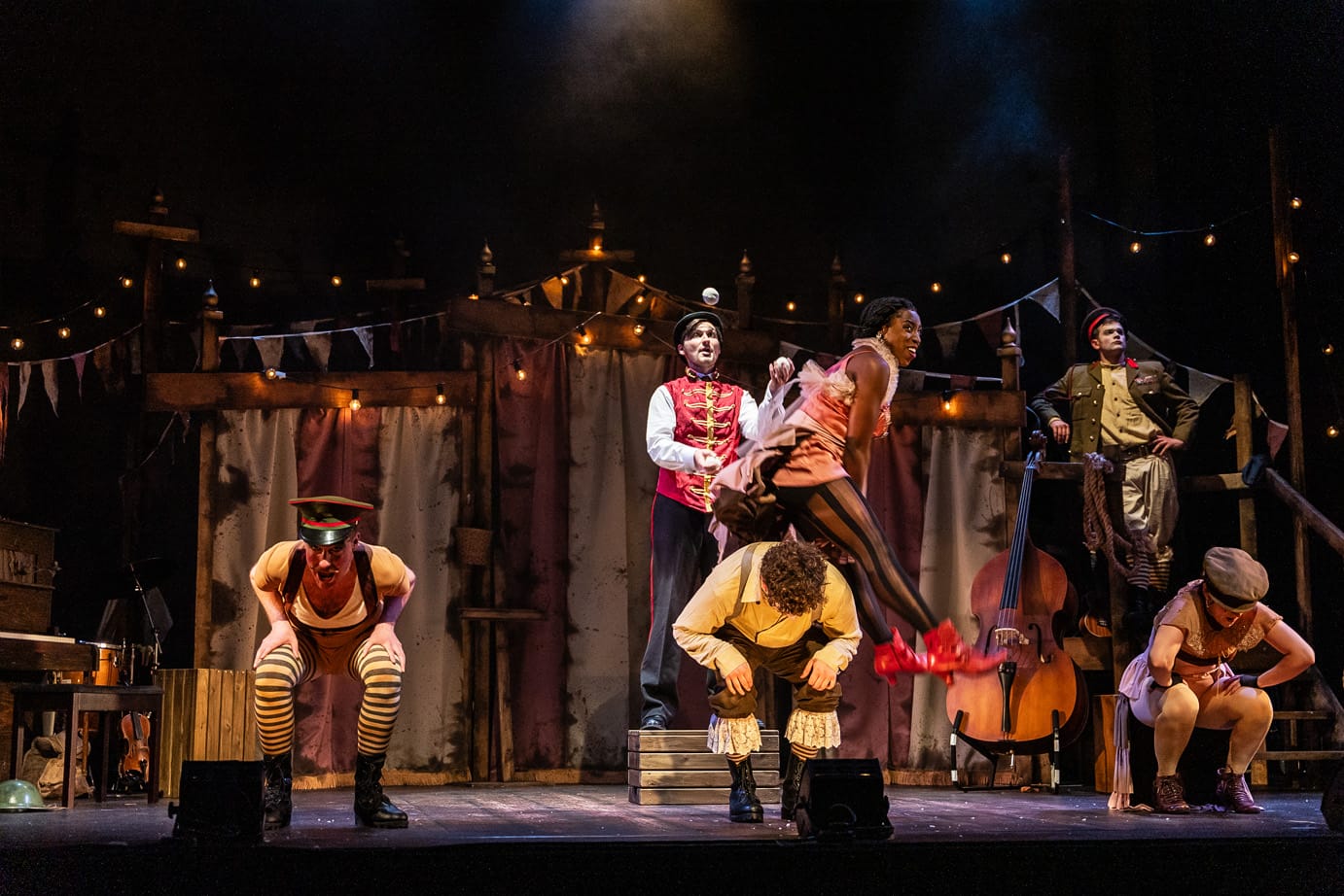 High quality production shot from a musical production of Oh What A Lovely War. The performers onstage wear vintage war-time garments with a burlesque twist. In the centre of the image, one performer is vaulting over another, grinning in mid air.