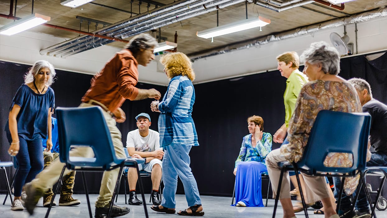 A busy workshop in a drama studio. Participants are mid-way through a movement exercise and are photographed in motion as they navigate the space.