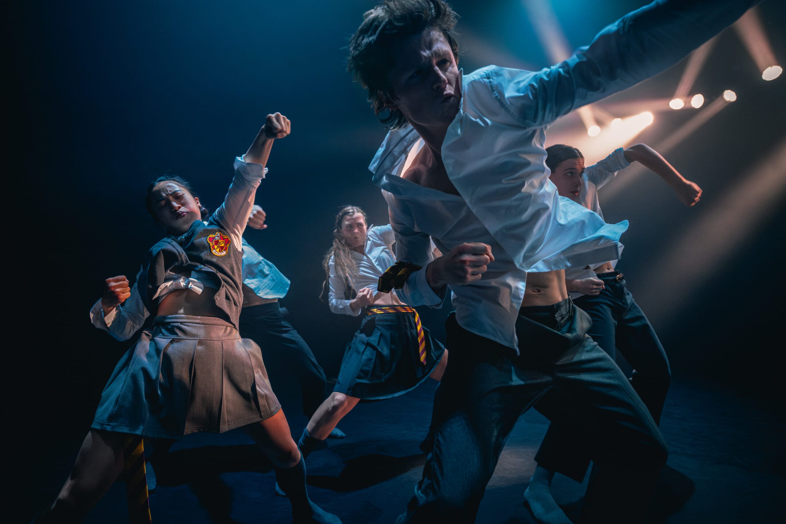 Several Shechter II dances are captured in dynamic poses in a motion shot. In the foreground, a male dancer punches the air with passion. All dancers wear contemporary, school uniform inspired costumes.