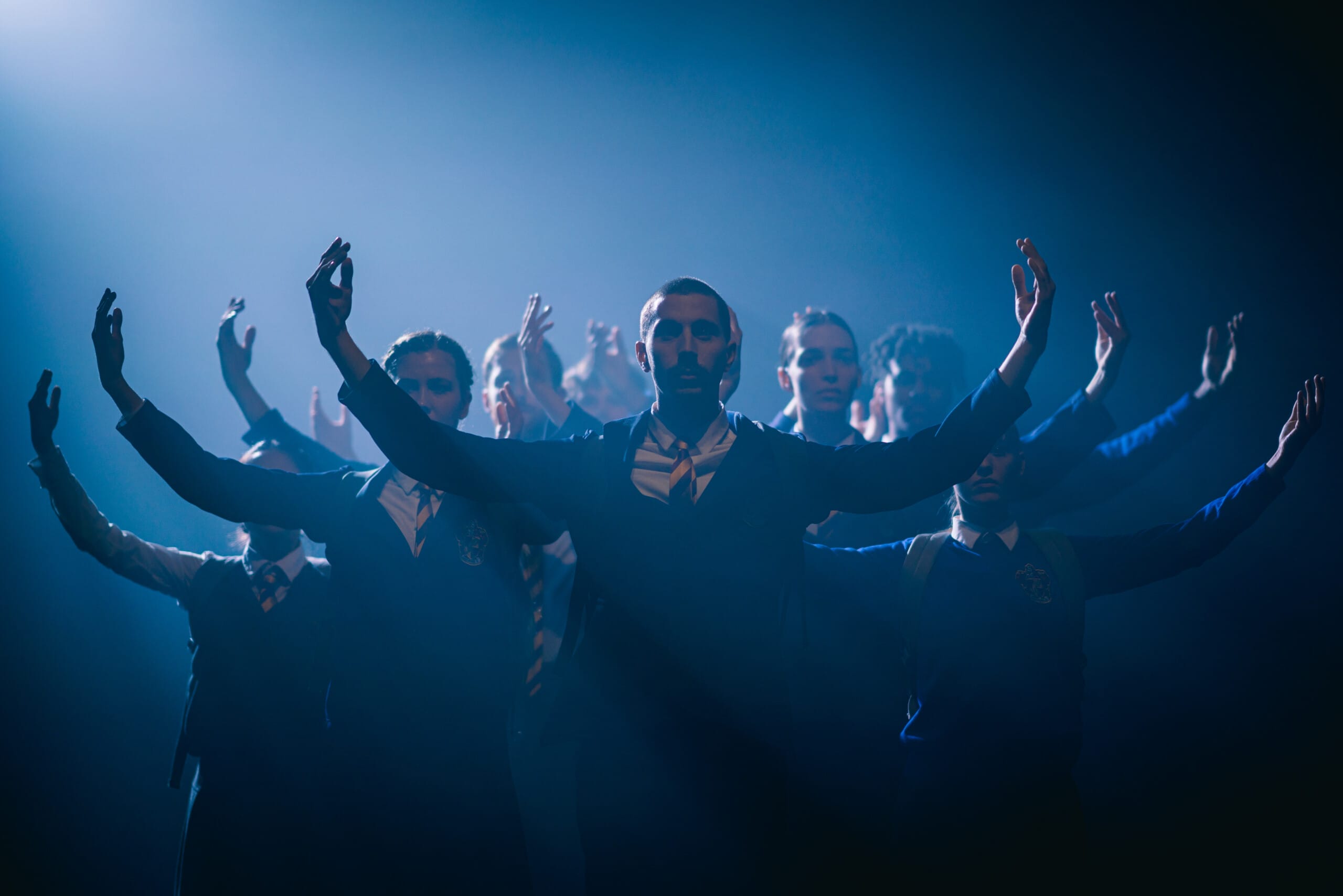 Dramatic blue lighting casts a shadow over the ensemble performers in Shechter II. All dancers pose with both arms raised in the air in a crescent position.