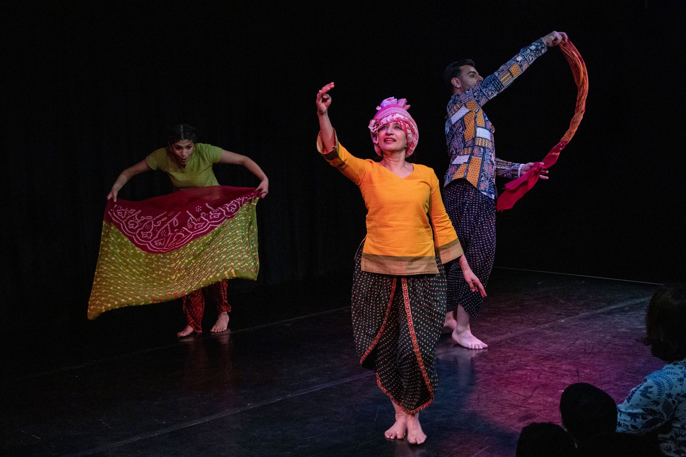 Onstage, three Desi performers are engaging in traditional Indian dance performance. The performer centre stage wears an orange shirt and pink headdress. The performers in the background are dancing with silk scarves.