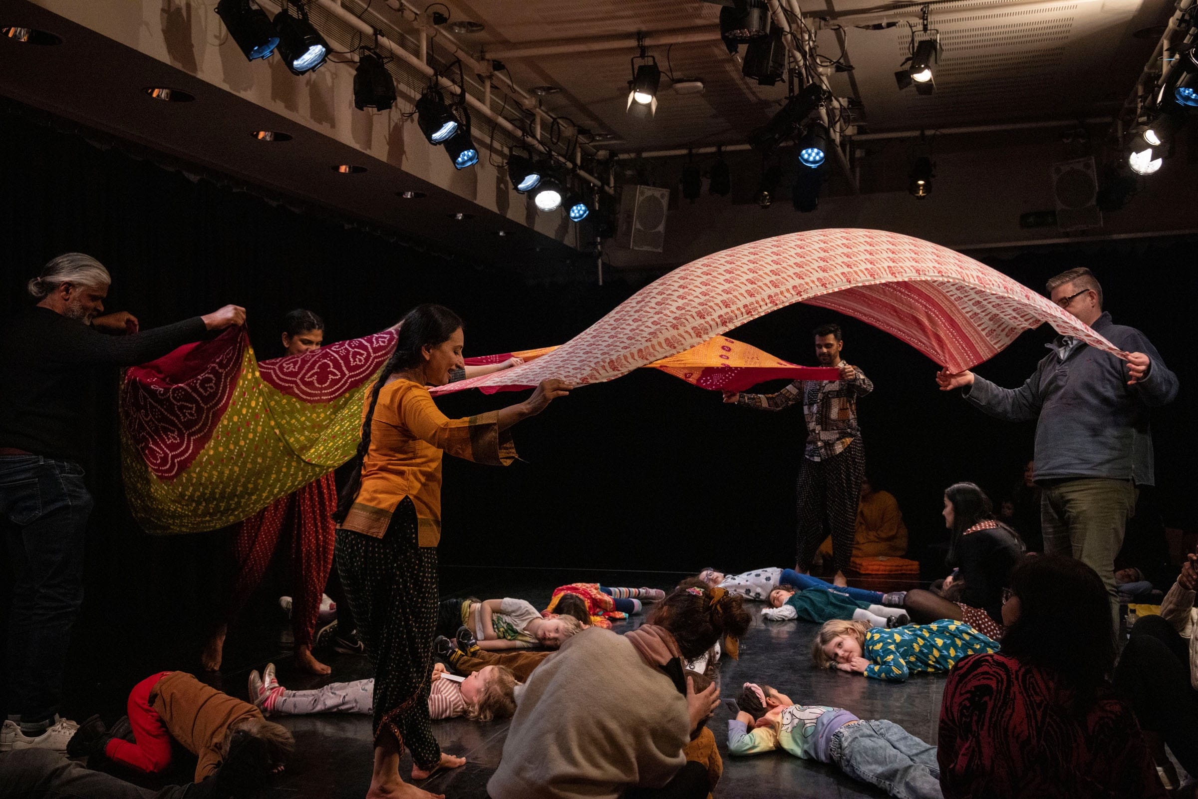 Performers throw billowing silk scarves into the air to create dynamic shapes. On the floor of the studio, children and their parents are laying on the floor, looking up at the silks as they engage in sensory play.