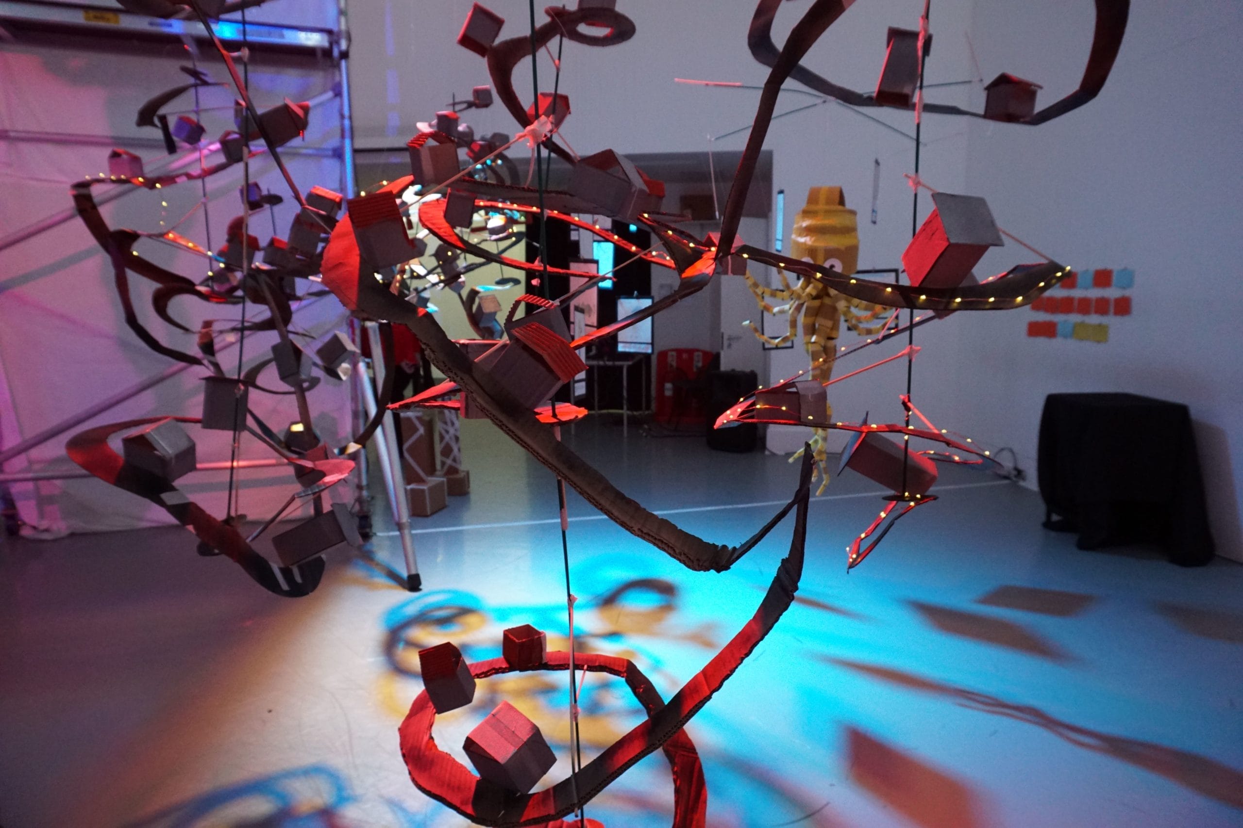 Wide shot of the exhibition space, where a futuristic world has been crafted out of cardboard and sustainable materials. Spiralling cardboard roads are hung from the ceilings with cardboard houses in the centre of the image. A cardboard octopus is visible in the background.