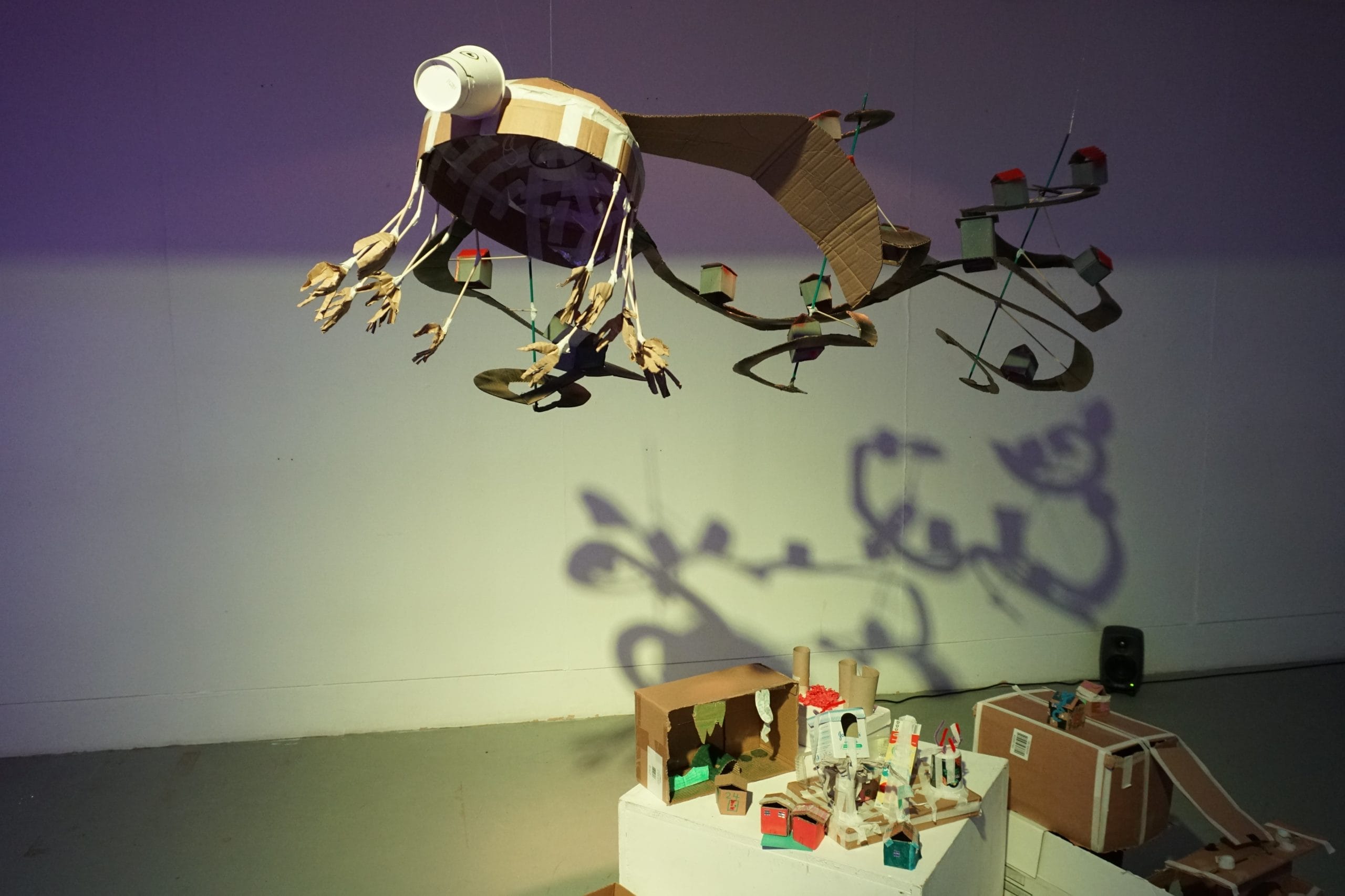 Cardboard houses and towns are constructed out of cardboard. Above them a large cardboard turtle-dragon is hung from the ceiling, decorated with other sustainable materials. The objects cause whimsical shadows to appear on the wall.