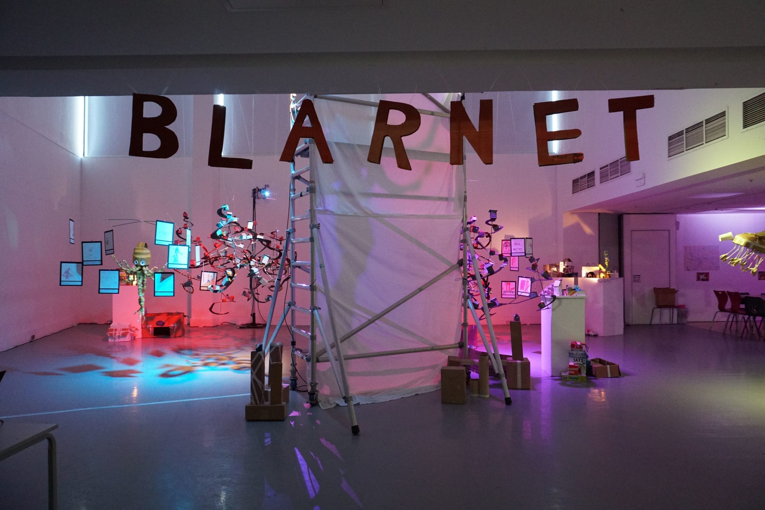Wide shot of the exhibition space, where a futuristic world has been crafted out of cardboard and sustainable materials. Hung from the ceiling in cardboard letters is the word BLARNET.