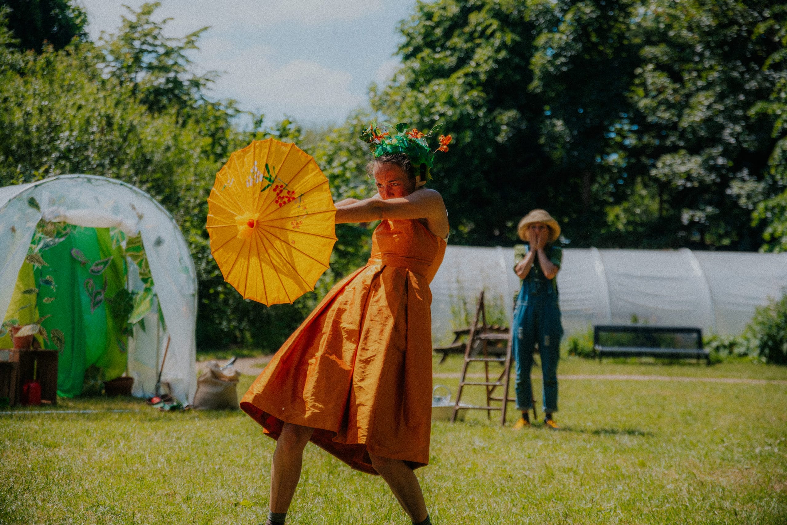 A green outdoor space. In the centre, the performer wears a vibrant golden dress, yellow parasol, and green floral headdress. In the background, another performer wears dungarees and a gardener's hat.