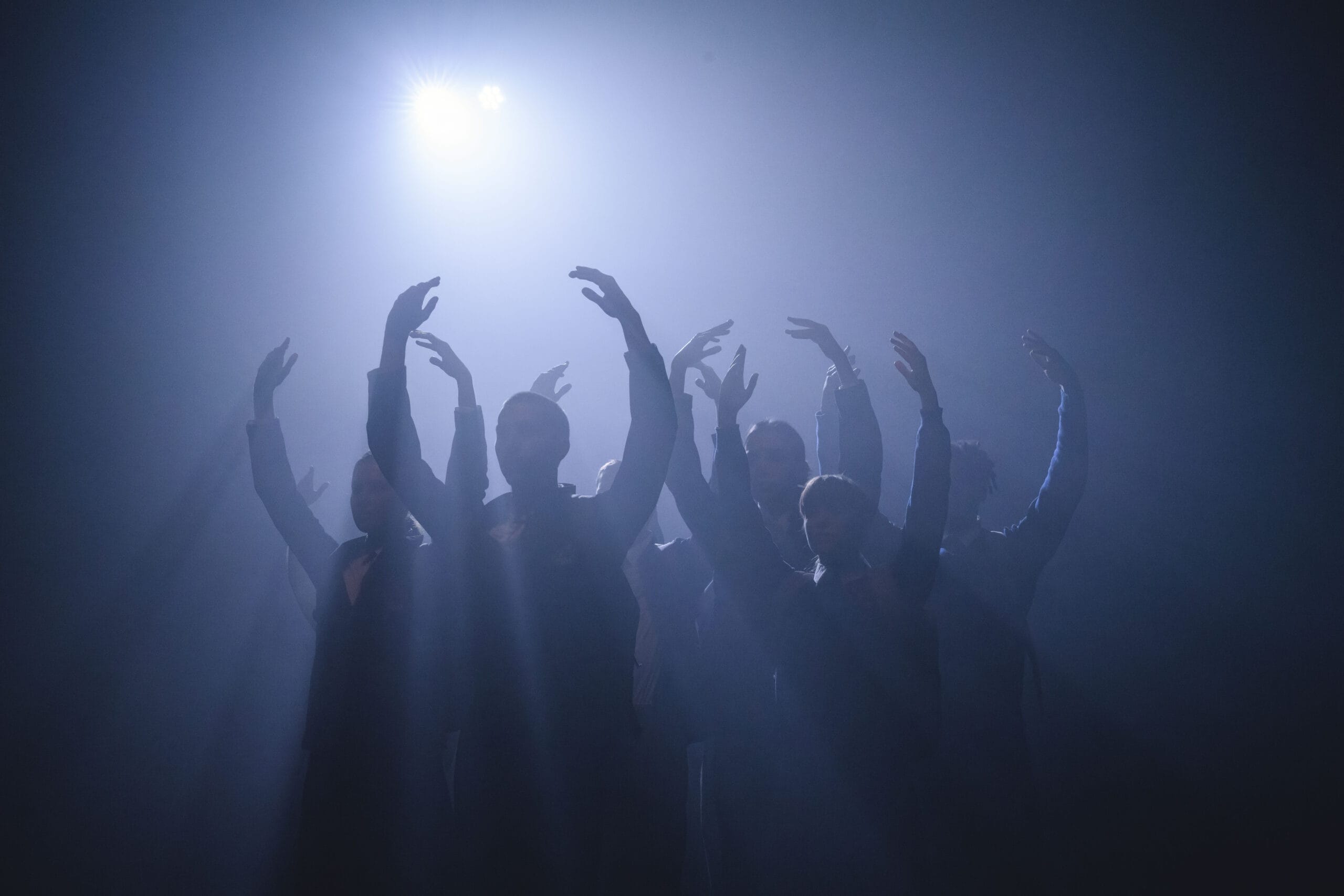 An ensemble of dancers stand in formation onstage with upstretched arms. A bold, blue light illuminates them from behind.