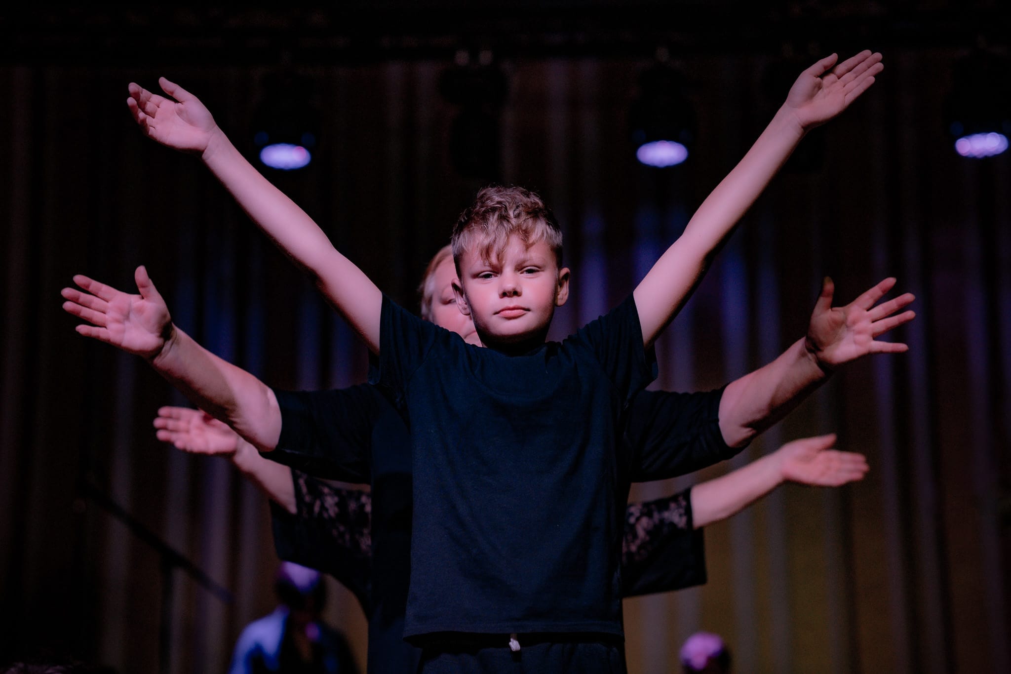 A group of dancers are standing one behind the other with their arms outstretched. Only the young dancer at the front of the line is fully visible.