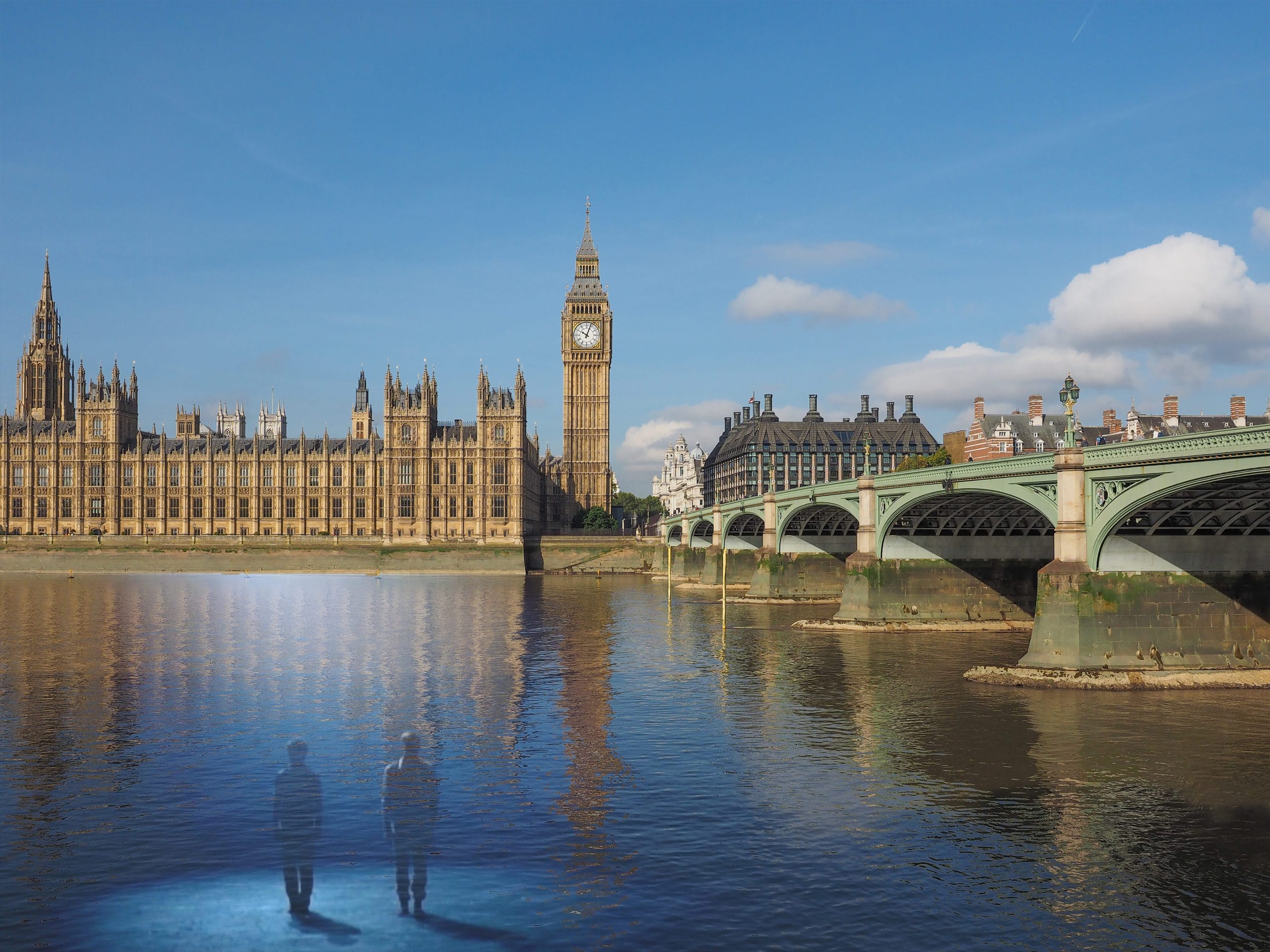 A landscape photograph of London landmarks including Big Ben, the houses of parliament, the River Thames and Westminster Bridge. In the left hand corner stand two silhouetted figures.