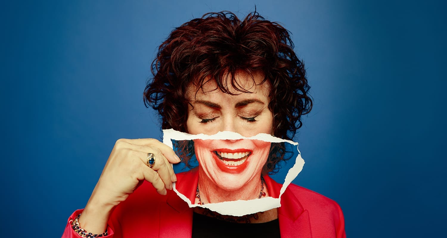 Ruby Wax poses against a dark blue background. Her eyes are closed, and she holds up a torn paper photograph of herself, smiling, positioned in front of the bottom half of her face. She is wearing a red cardigan and red lipstick.