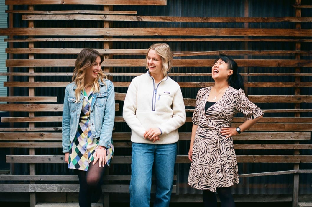 Three women stand in front of a wall made of wooden planks. They are smiling and laughing.