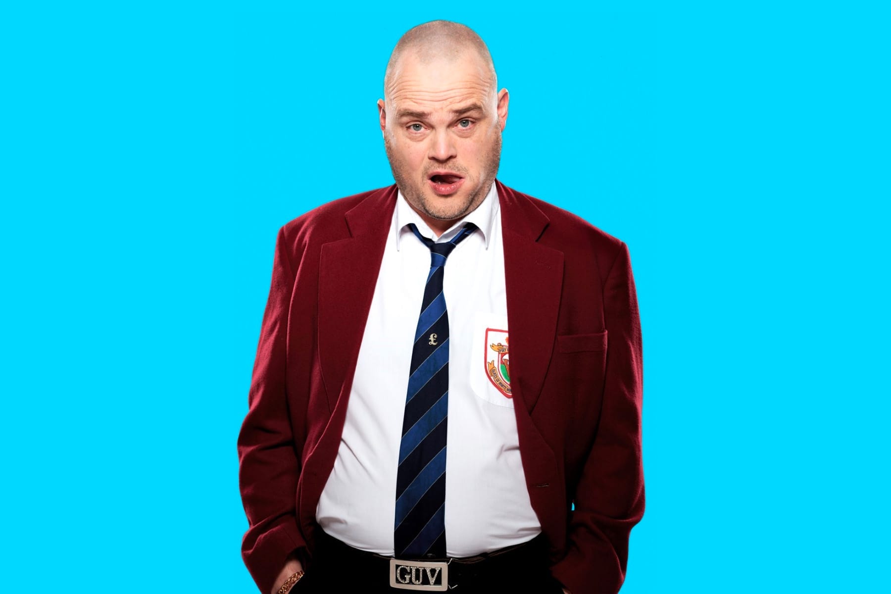 Comedian Al Murray poses in a maroon blazer and striped school tie. His belt is bedazzled with the word 'GUV'. Behind him is a vibrant blue background.