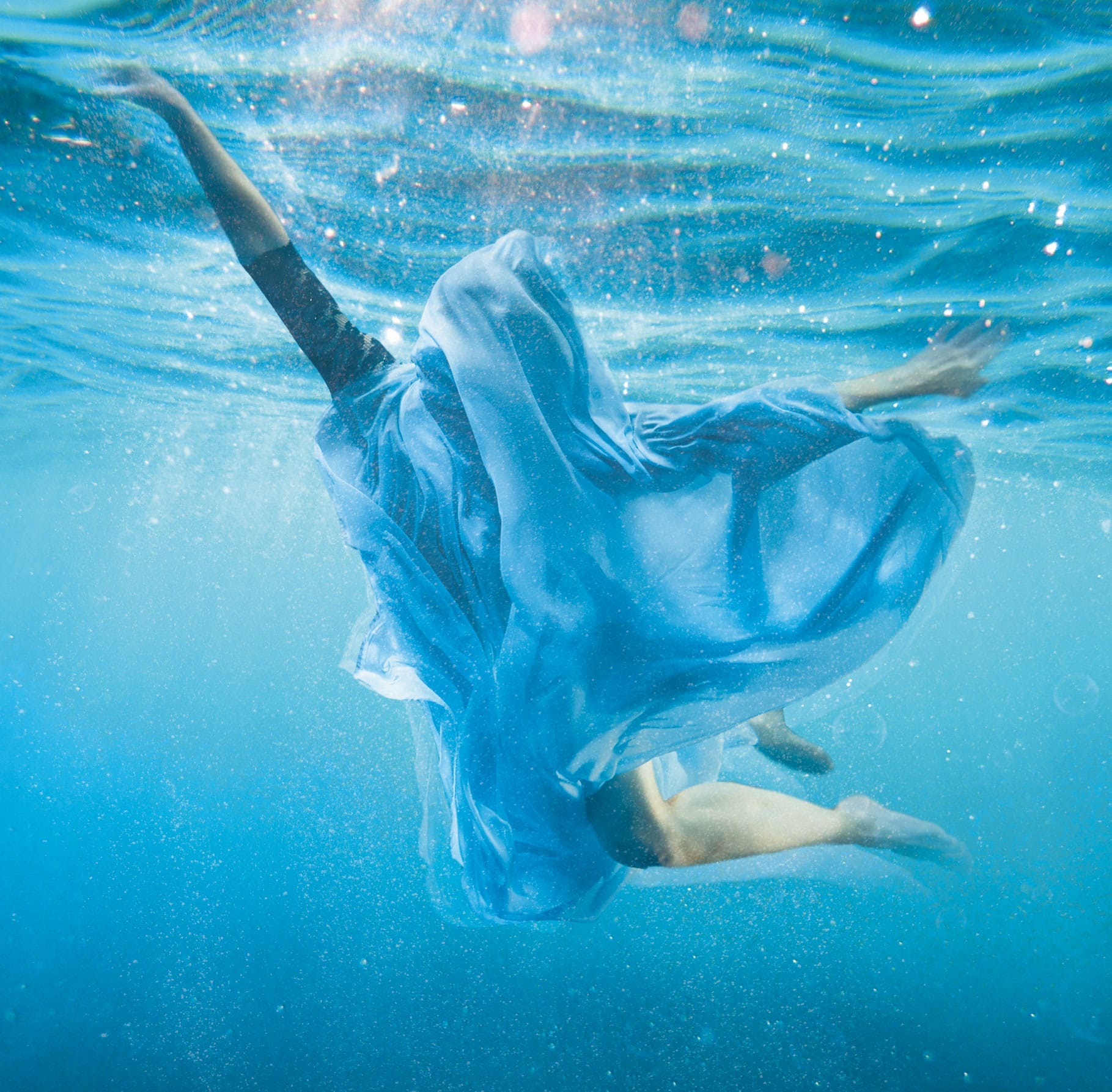 A dancer poses underwater in a clear blue ocean setting, with light streaming down from the surface. The dancer is covered entirely in flowing blue fabric, almost allowing them to blend in with the scenic waves.