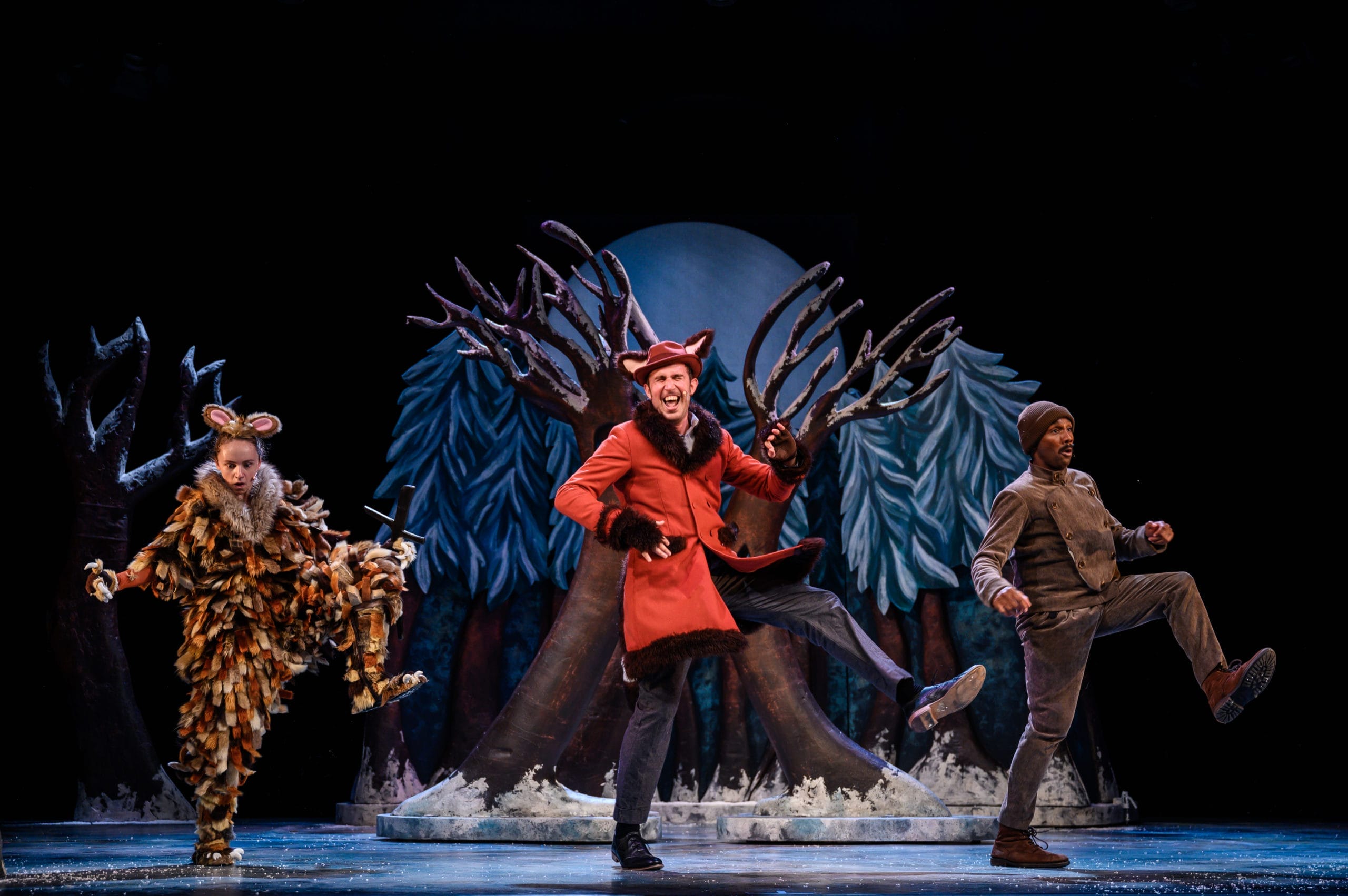 Production shot from The Gruffalo's Child artsdepot! The Gruffalo's Child, Fox and Mouse are dancing with their right legs kicking in the air. Behind them is snowy woodland scenery.