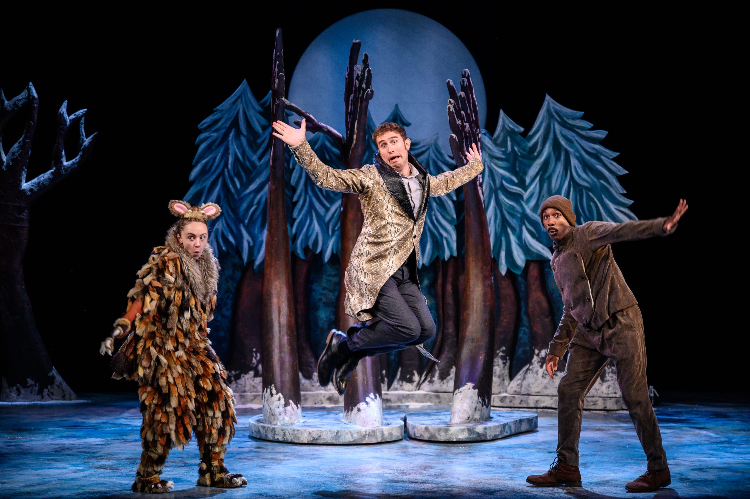 Production shot from The Gruffalo's Child artsdepot! The Gruffalo's Child and Mouse look out to the audience with Snake in the middle. Snake is leaping in the air with a silly expression, wearing a long snakeskin coat.