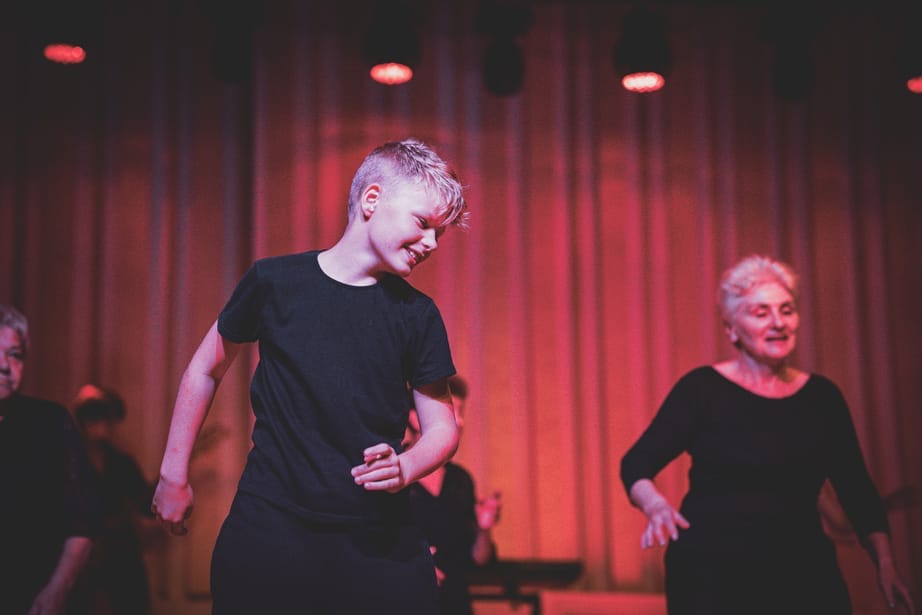 A teenage boy and an older woman dance side by side on stage. They are both wearing black outfits and smiling.
