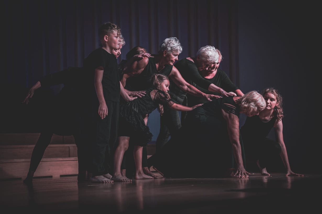 A group of dancers of all ages, pictured on stage. A senior woman is leaning over, other dancers reach out to her.
