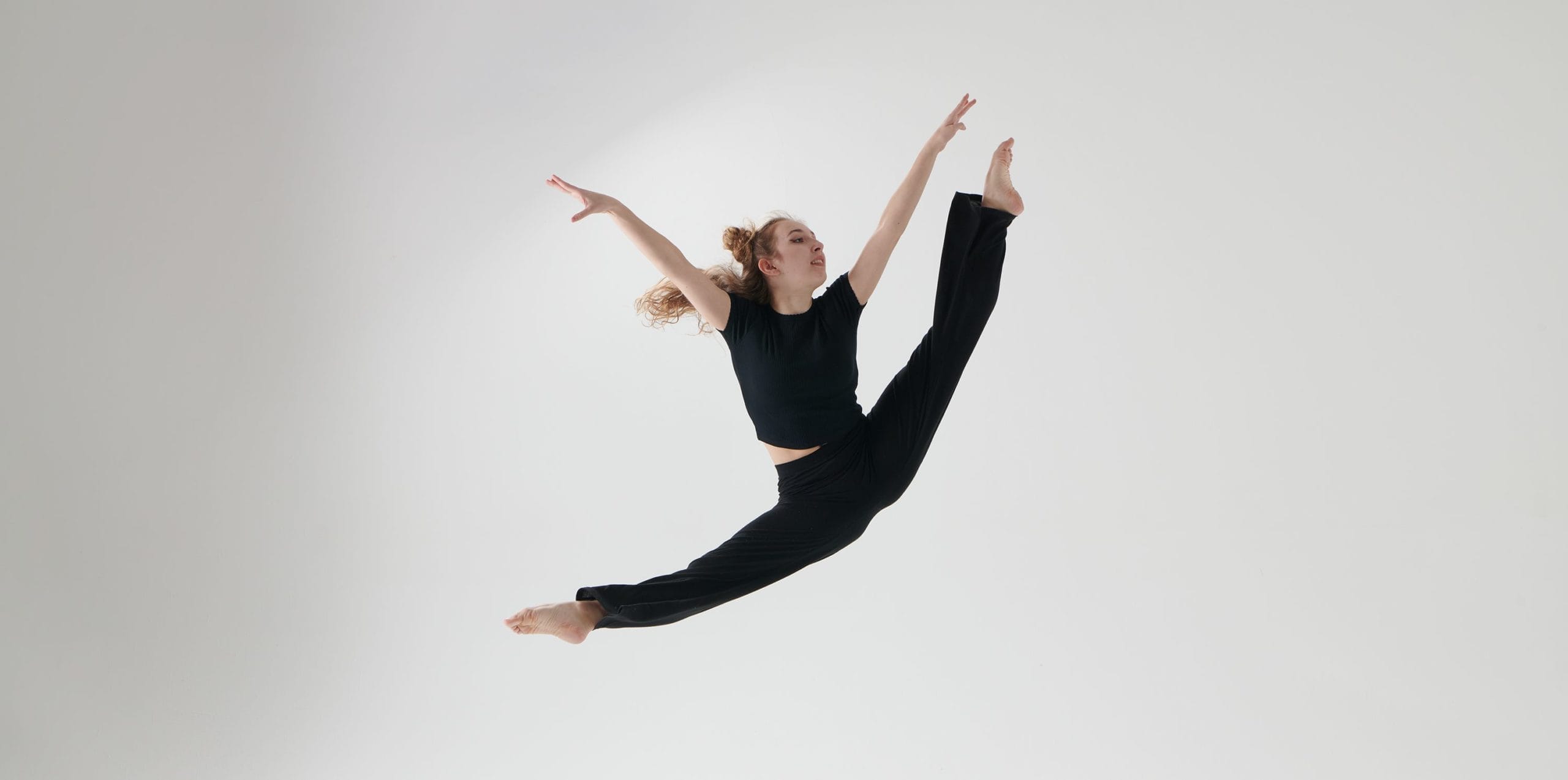 A blonde female dancer is photographed against a white background. She is leaping into an impressive oversplit, with pointed feet and arms in the air. She is wearing a black t-shirt and black trousers.