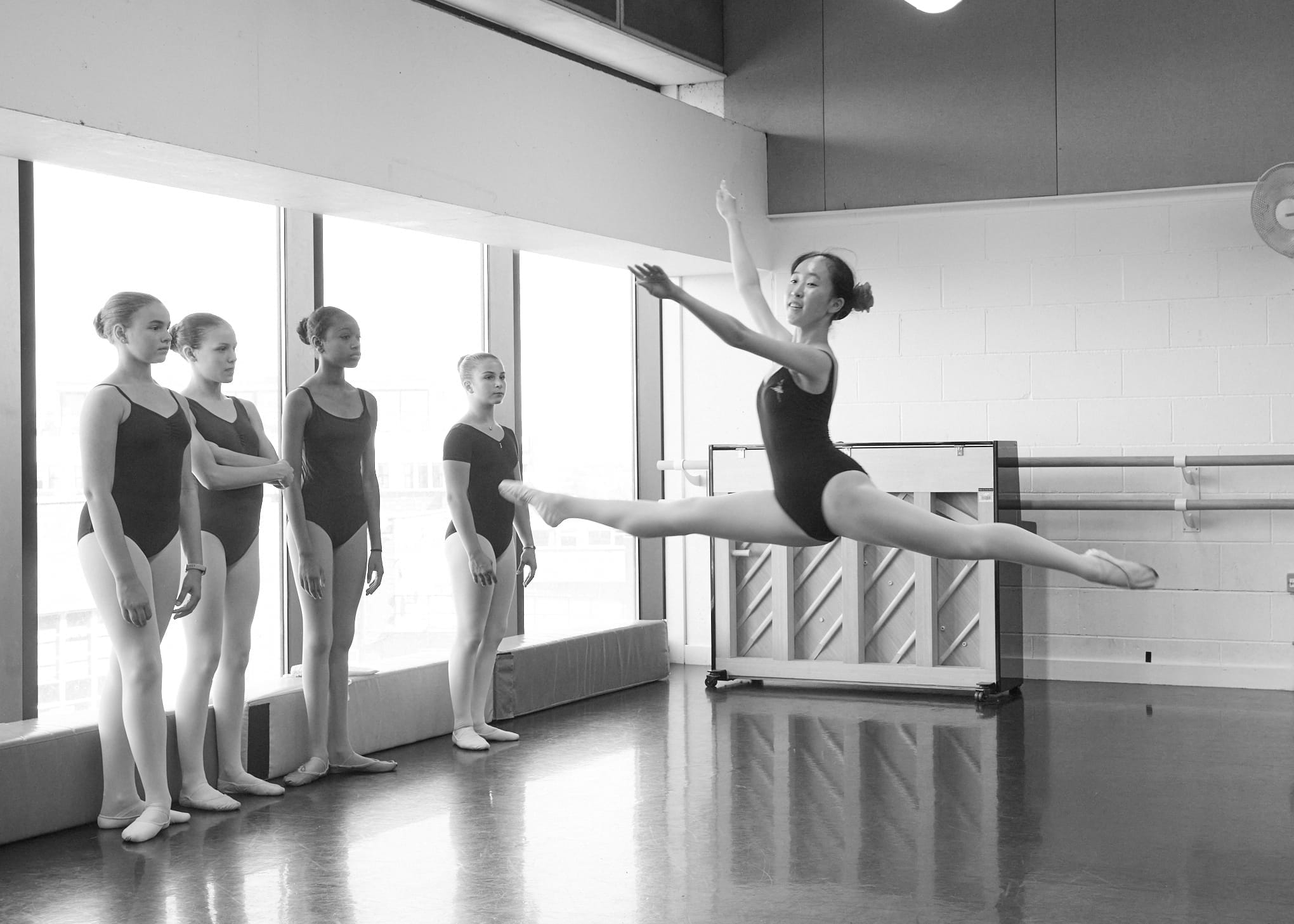 A black and white photo of a ballet studio, where a diverse group of dancers in black leotards are gathered. Centre stage, a featured dancer is leaping in an impressive arabesque.