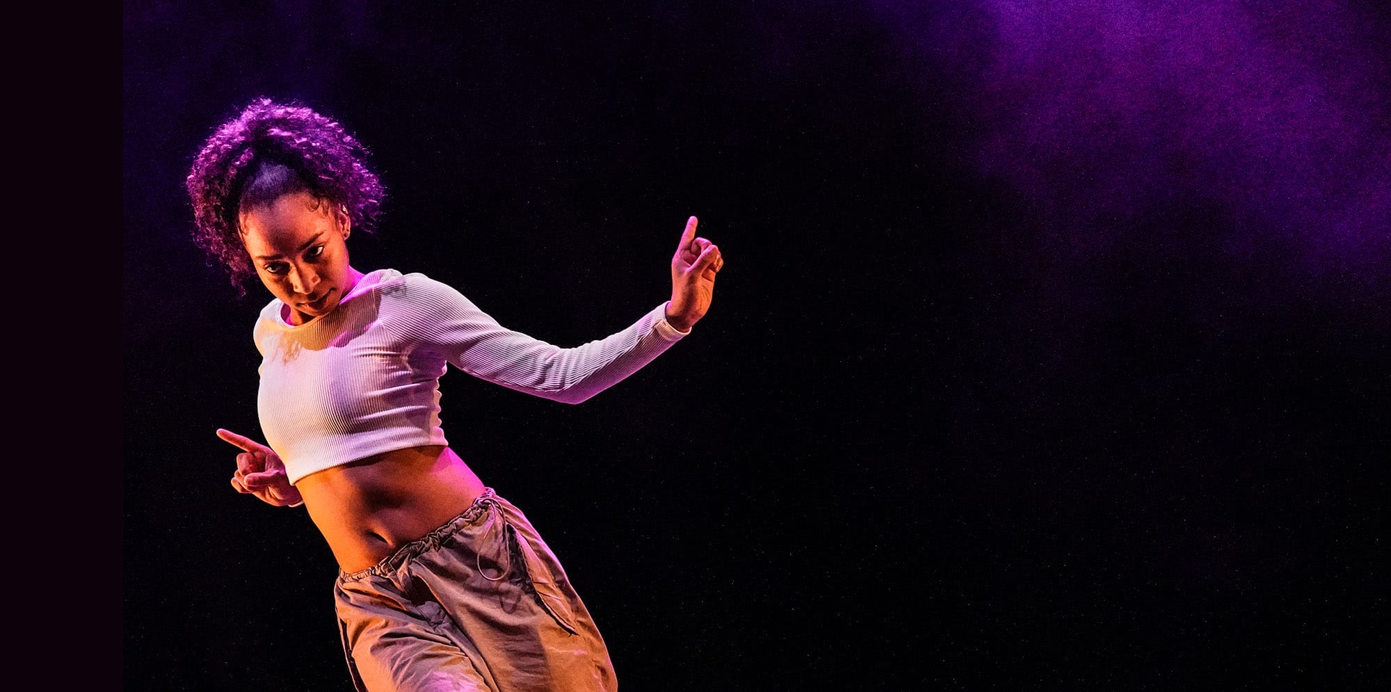 A female dancer is featured onstage. She has curly brown hair in an updo, a white long sleeve crop top and cargo pants. She is illuminated by purple stage lighting.