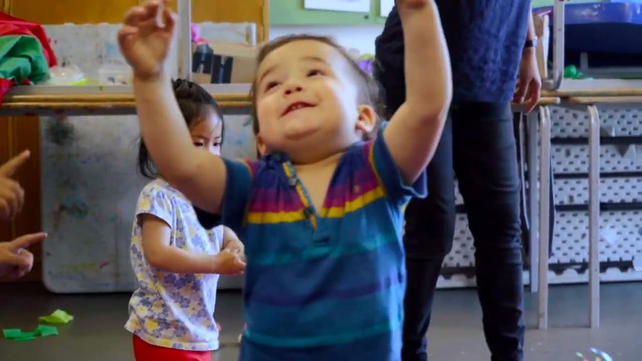 A happy toddler is featured with his arms in the air and a big smile. He is wearing a colourful stripy shirt. Another toddler and their adult are visible in the background.