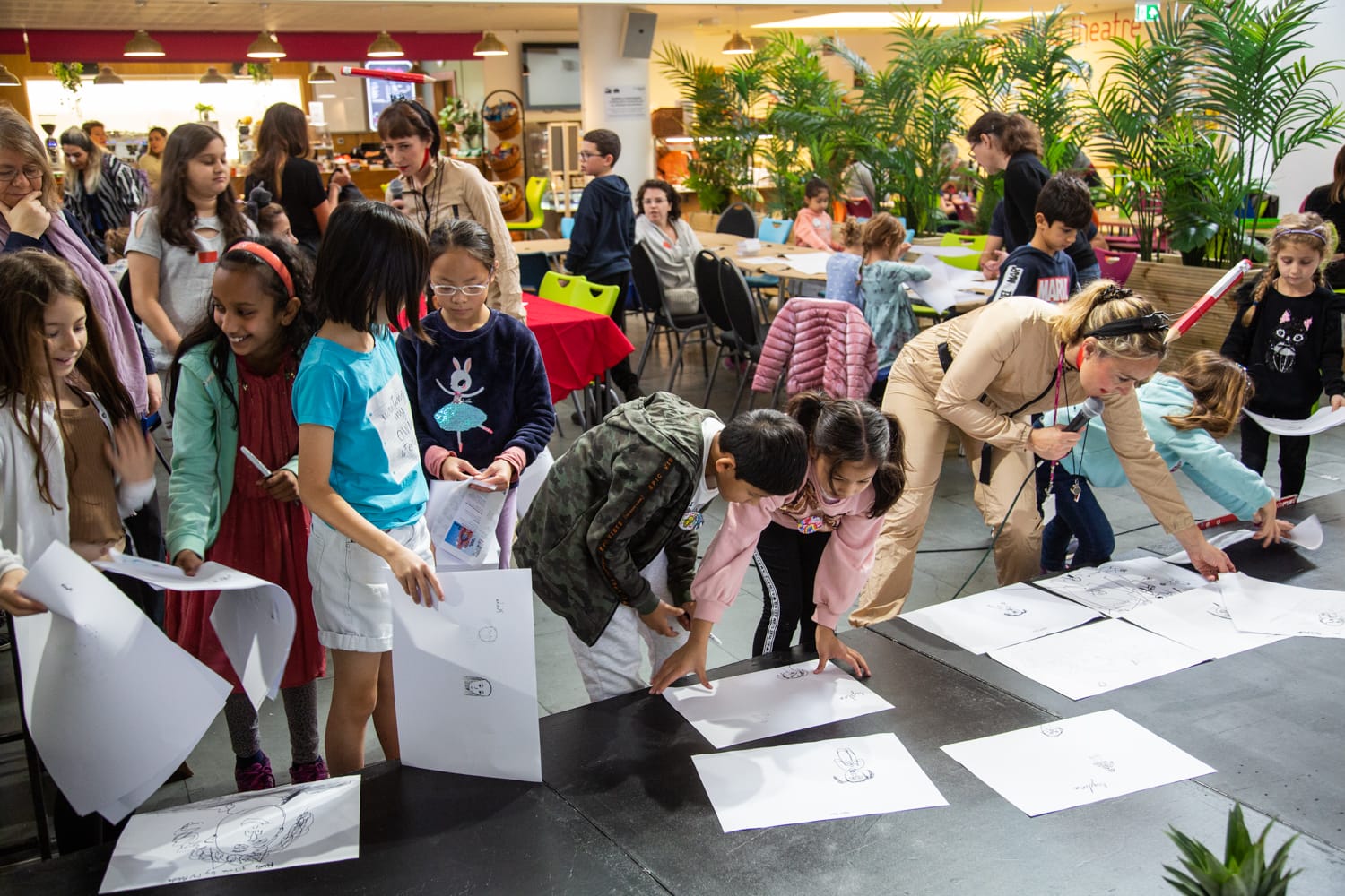 A diverse group of children and parents are gathered in the artsdepot cafe, smiling, chatting, and working on pencil drawings. At the front, several children are holding large a3 sheets of paper.