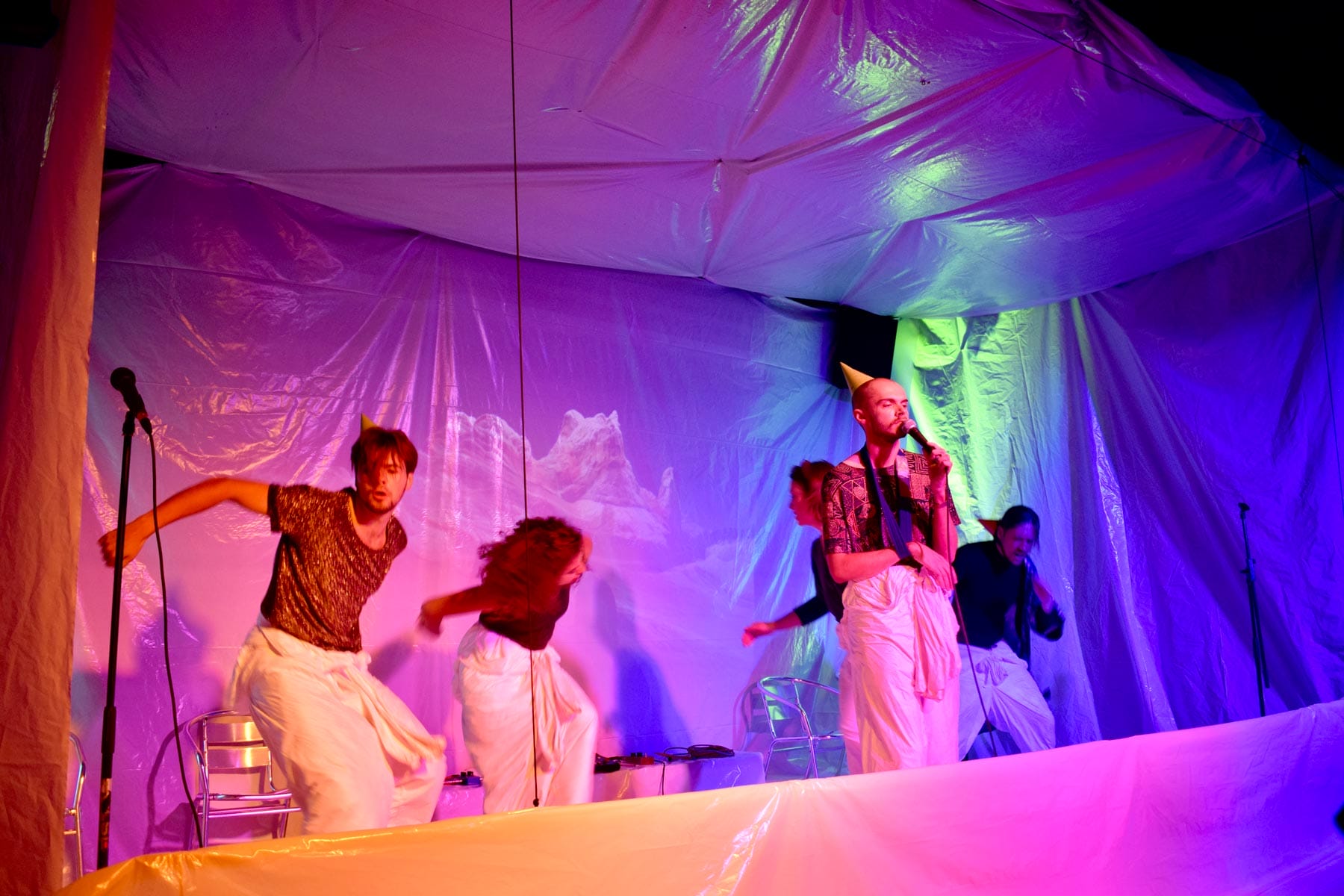 A group of performers on a stage covered in white plastic sheeting, they wear white boiler suits tied at the waist. One wears a sling and a party hat, the others are dancing energetically.