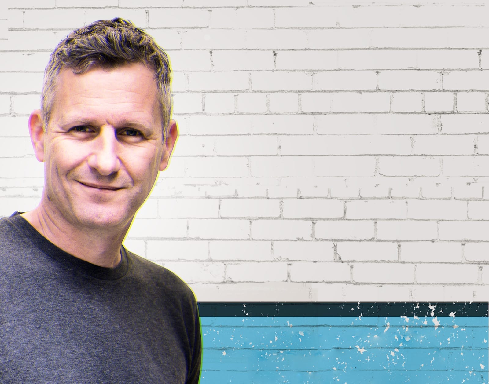 Comedian Adam Hills poses to the left. He has short grey-brown hair and is wearing a grey t-shirt. The background is a white brick wall with a blue stripe at the bottom.