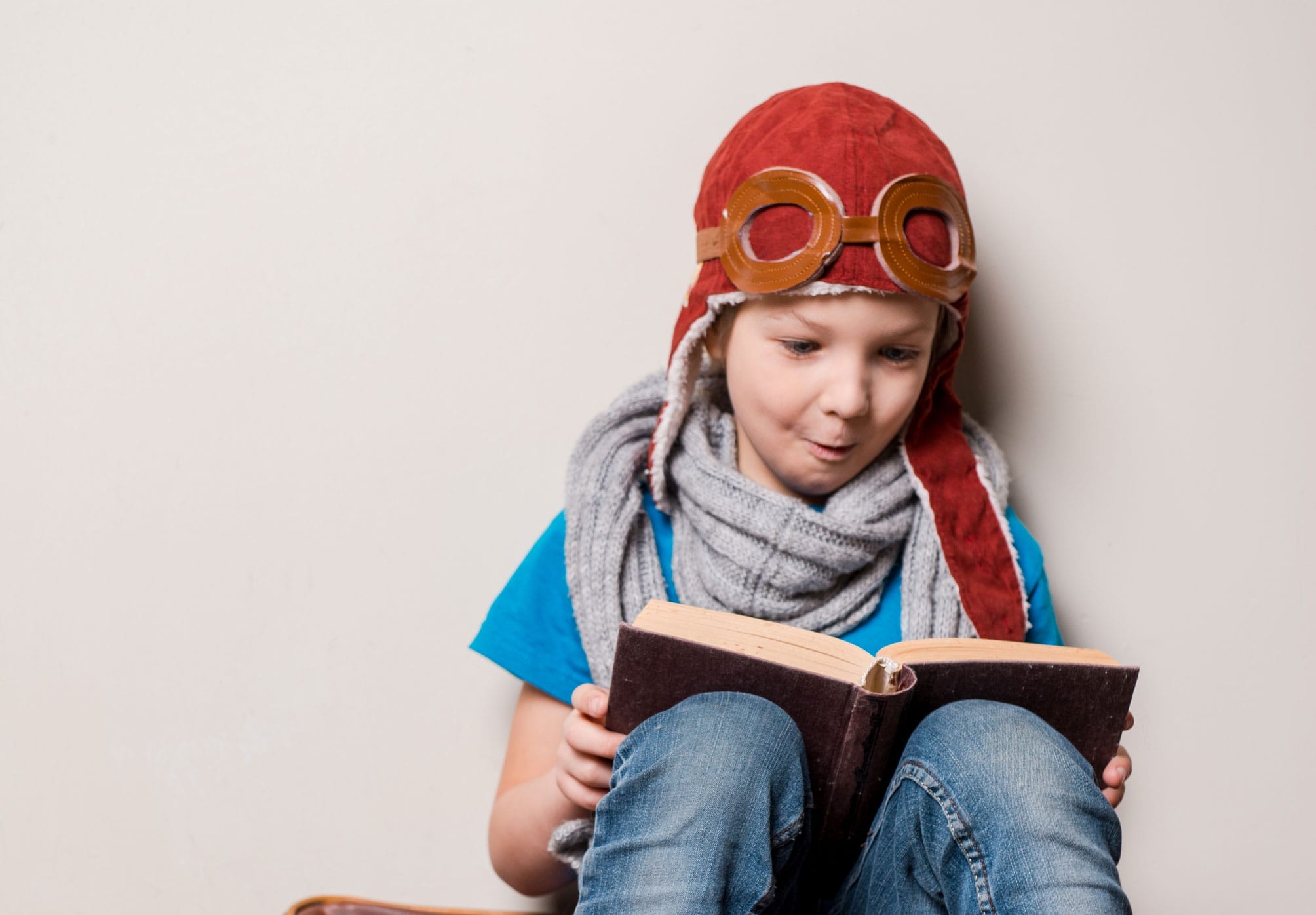 A young boy in a red flying helmet sits on a leather suitcase reading a book.