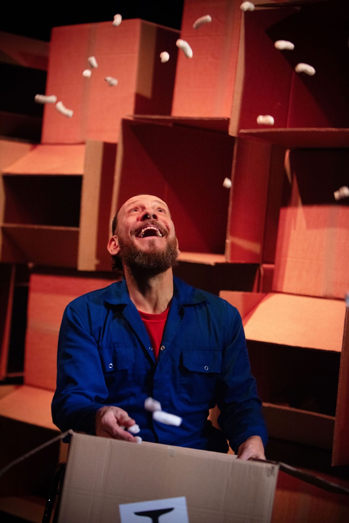 Daryl, a white man with a beard in light blue overalls, looks upwards as white packing peanuts rain down on him. He is surrounded by cardboard boxes