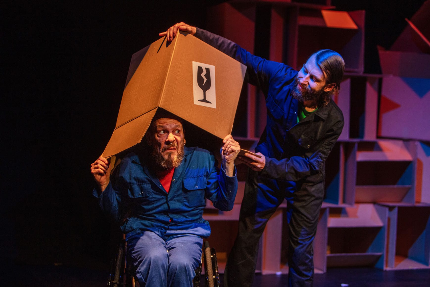 Daryl, a white man with a beard in light blue overalls, is sat in his wheelchair peering out from a cardboard box on his head. Jon, a white person with a beard wearing dark blue overalls is lifting the box of Daryl’s head. In the background is a wall of cardboard boxes