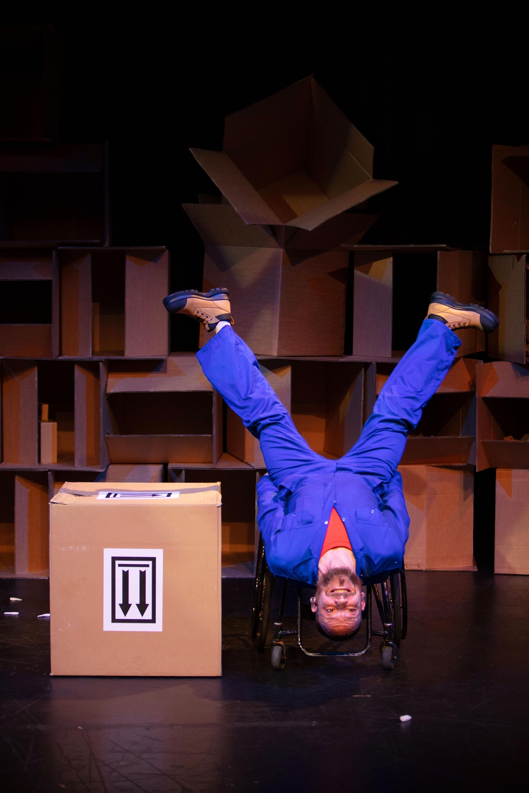 Daryl, a white man with a beard in light blue overalls sits upside down in his wheelchair with his legs in the air. In the background is a wall of cardboard boxes