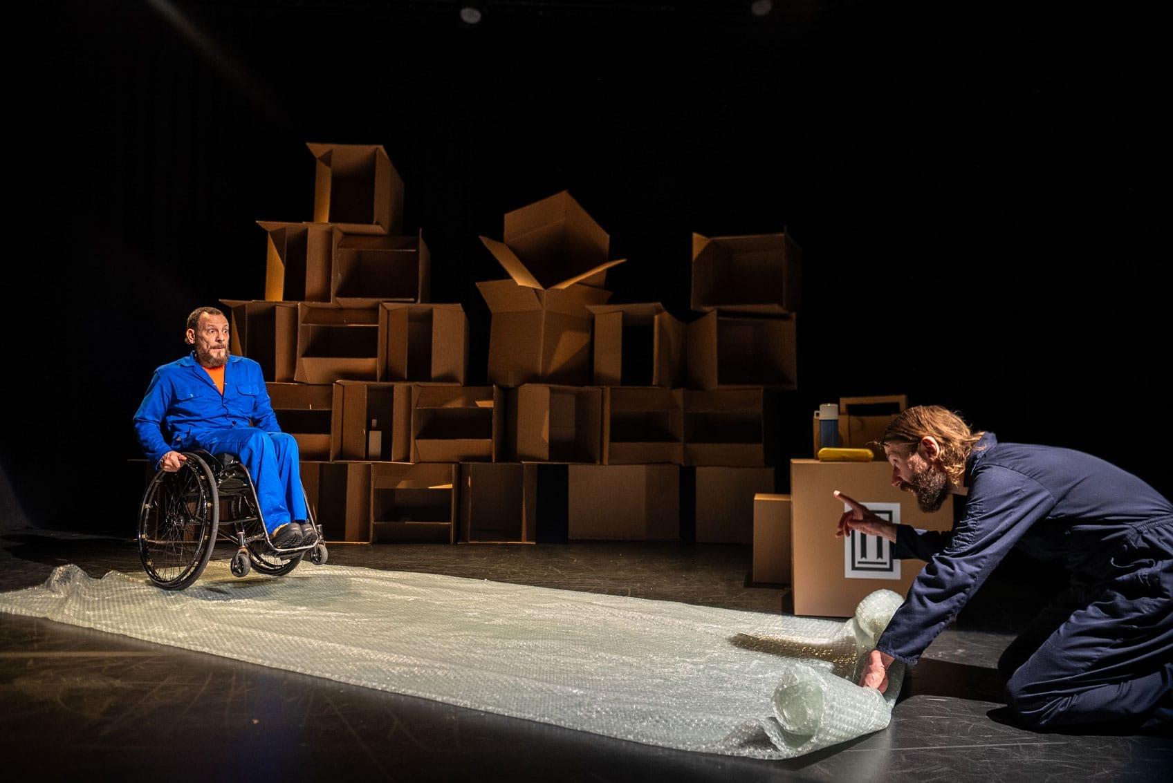 On the floor is a large piece of bubble wrap layer out. At one end Daryl, a white man with a beard in light blue overalls sits in his wheelchair on top of it. in bubble wrap. Jon, a white person with a beard wearing dark blue overalls is at the other end gesturing Daryl to stop. In the background is a wall of cardboard boxes