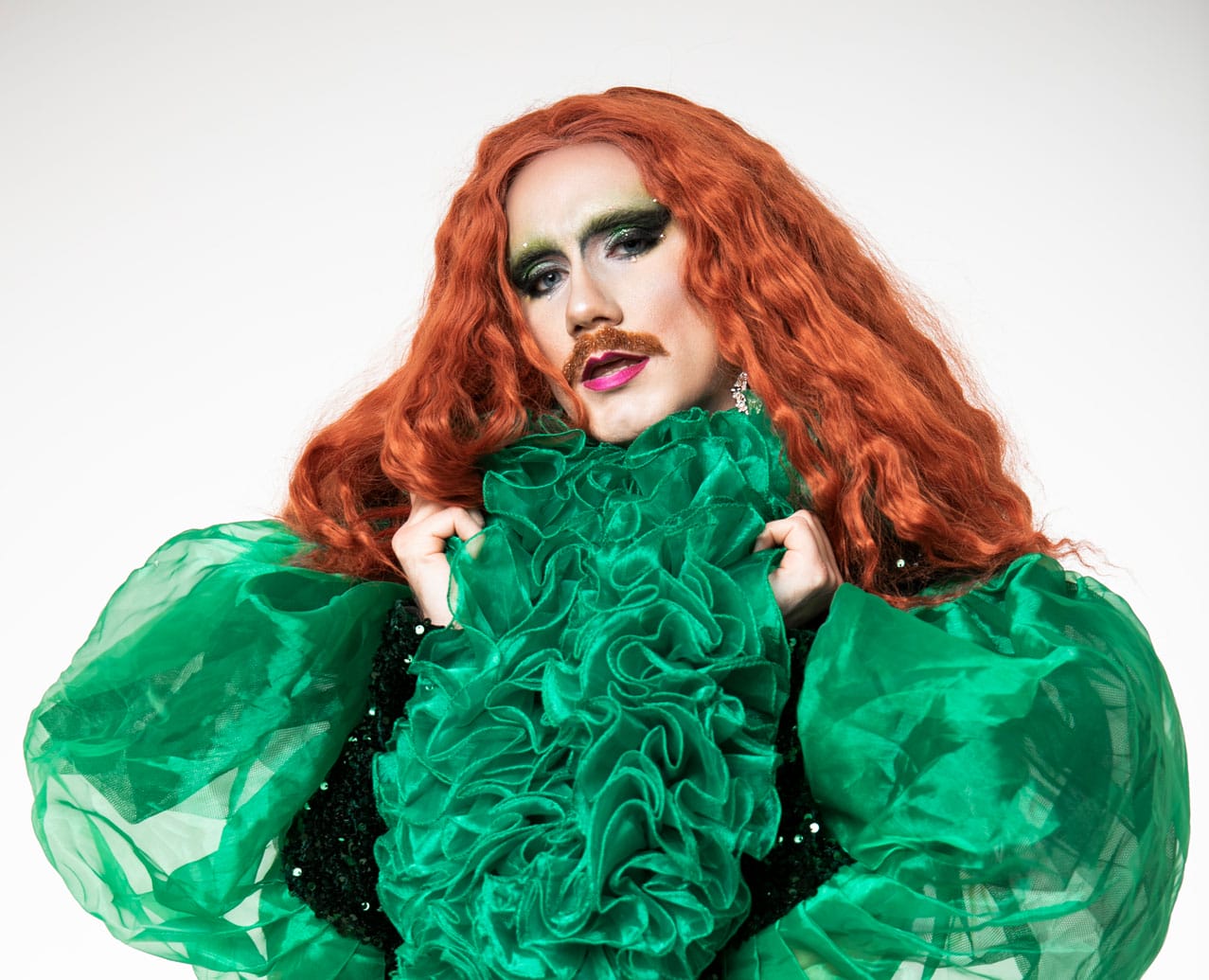 Carrot, a drag artist with long red hair is looking straight at the camera, they are wearing a green outfit with a lot of ruffles around the neck and down the front and large puffed sleeves.