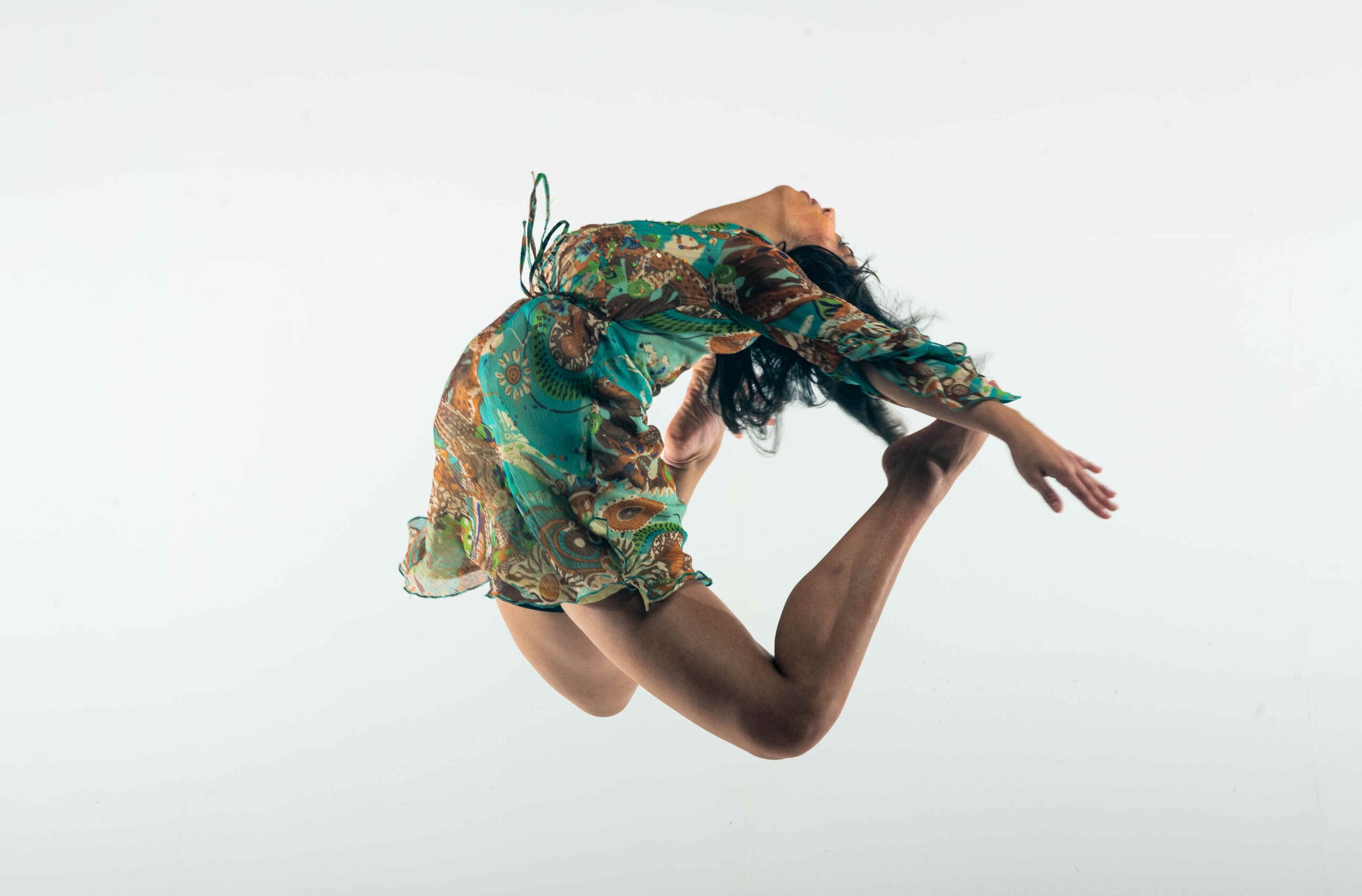Against a white background, a female dancer is photographed mid jump, displaying impressive flexibility. She has an arched back, with her arms thrust backwards, feet touching her hair. She is wearing a floral turquoise dress.