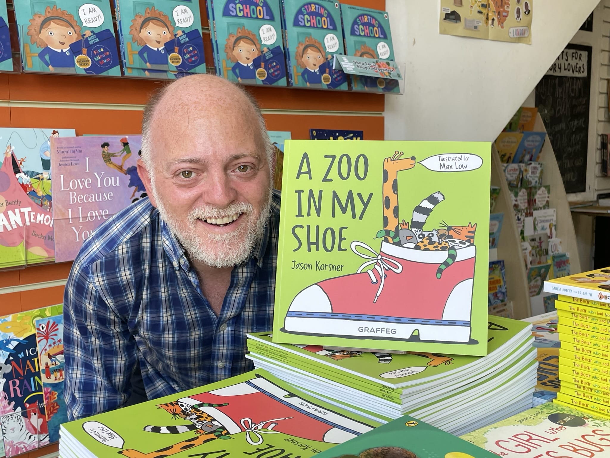 Children's author Jason Korsner poses with his book, A Zoo in my Shoe! The book is vibrant green with a red converse and giraffe on the front cover. Jason has grey hair, is smiling and wearing a blue checked shirt.