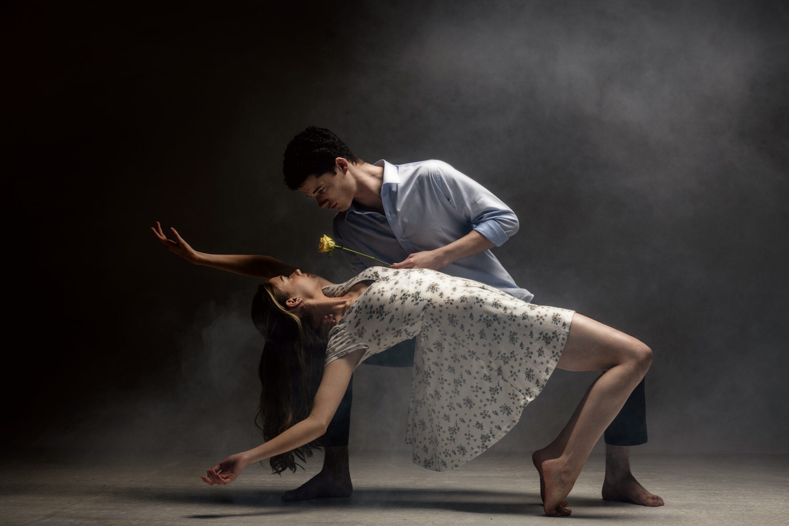 Against an atmospheric grey background, a male and female dancer engage in a duet. The female dancer is arching backwards in a romantic pose. The male dancer holds a yellow rose.
