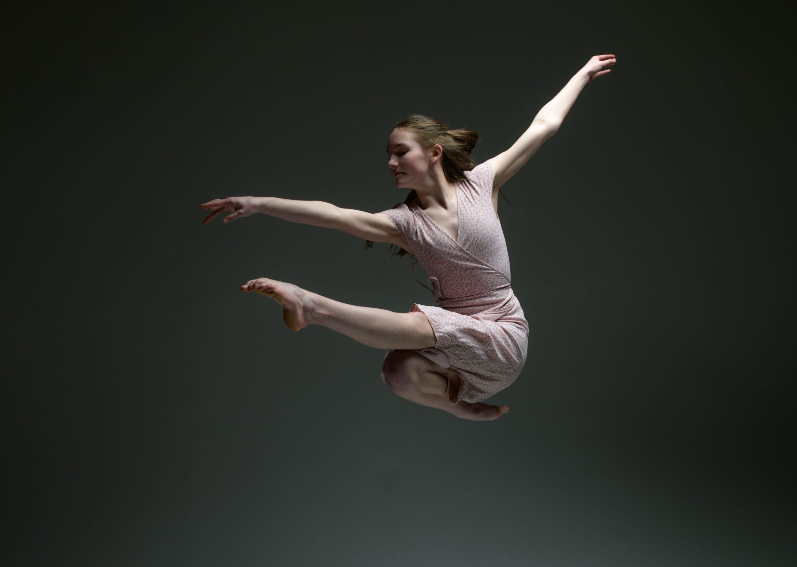 Against a dark grey background, a female dancer is photographed mid leap. One leg is extended out, with the other tucked beneath. Dancer wears a pale pink linen dress.